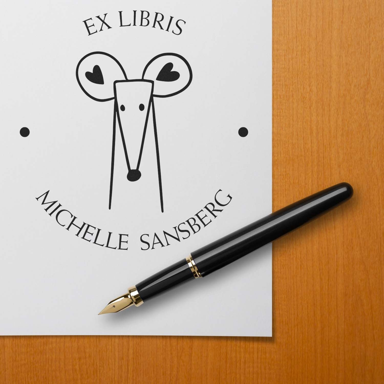 DazzleDash Cartoon Animal Pre-Inked Ex Libris Library Stamp featuring a stylized mouse design on paper with "Michelle Sansberg" text. A Custom Address Stamp is shown next to a black fountain pen on a wooden surface.