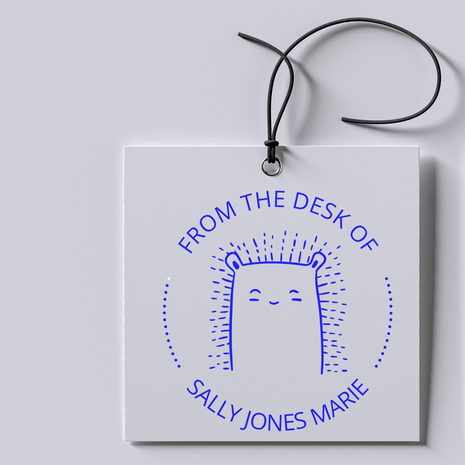 A FizzleFluff cartoon animal pre-inked book stamp on a white tag with From the Desk of Sally Jones Marie in blue. The personalized mailing stamper features a cute, smiling creature with spiky hair, hanging by a black string.