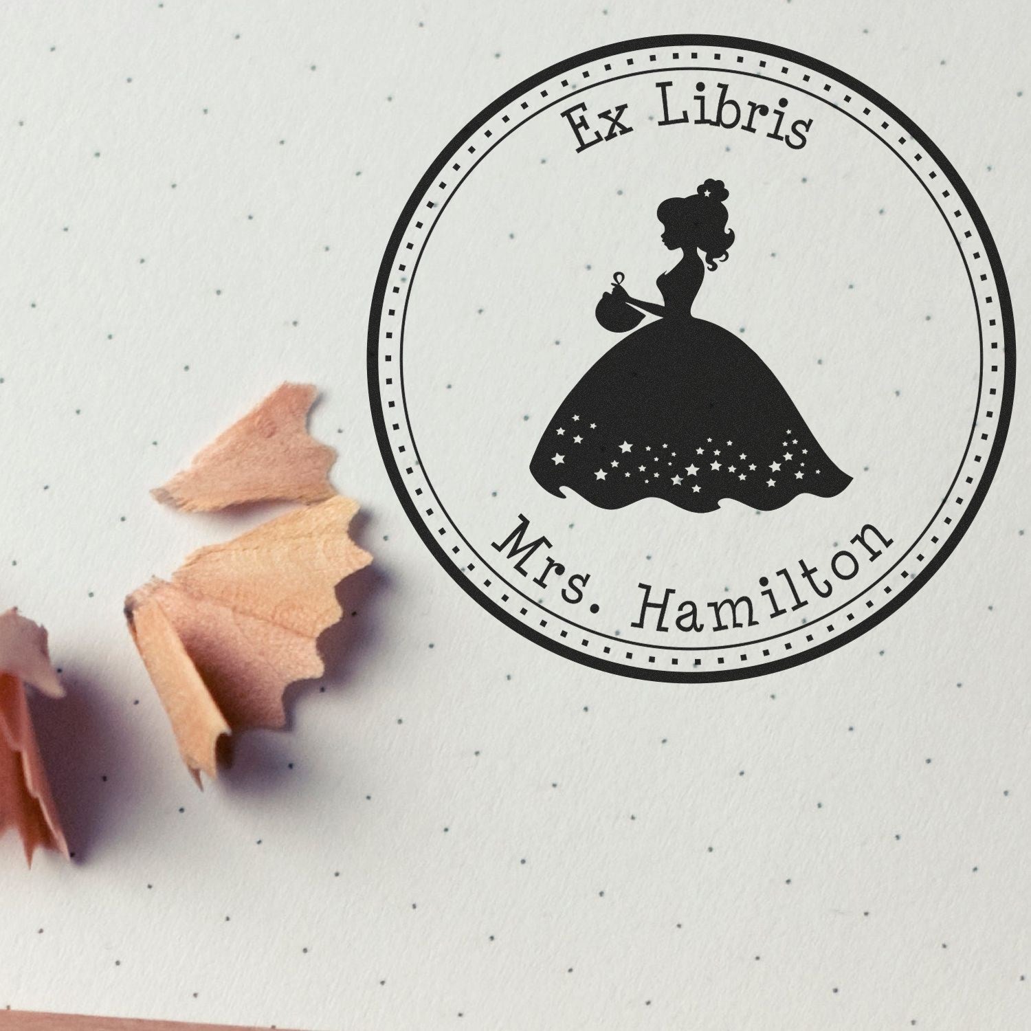 Black round Ex Libris stamp seal for Mrs. Hamilton with silhouette in gown on white dotted paper and pink shavings.