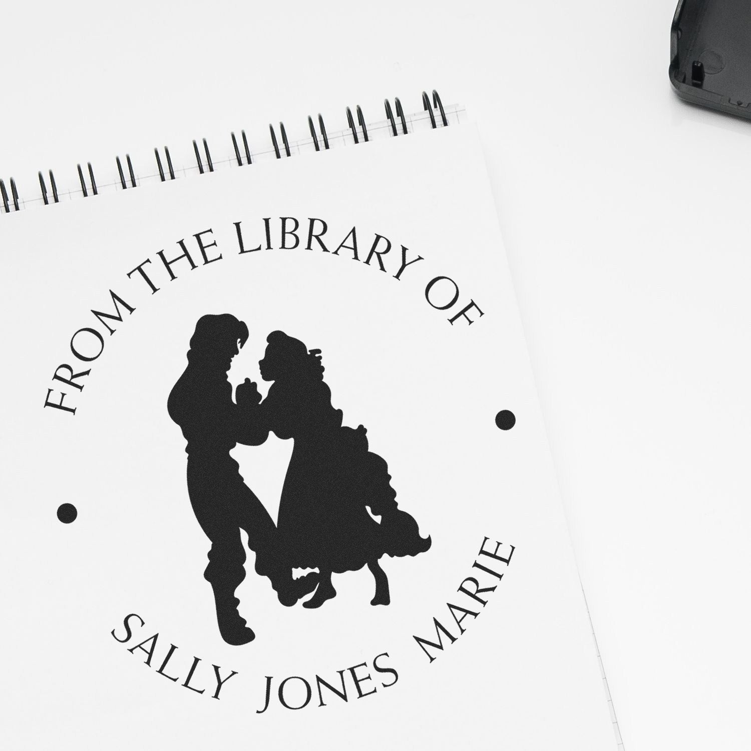 Black custom self-inking library stamp and seal with silhouette and curved text on white notebook paper.