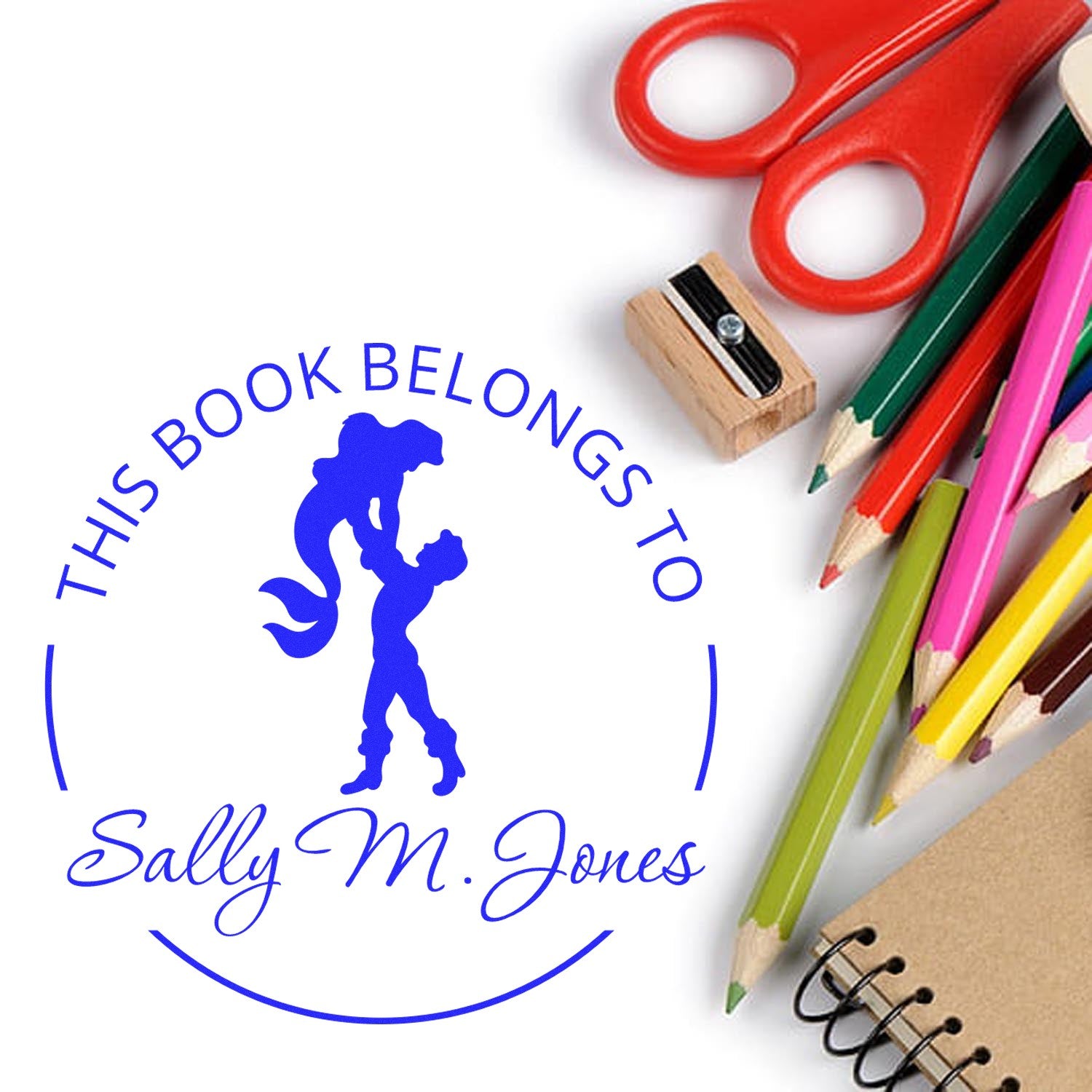 Blue custom book stamp seal with mermaid and Sally M. Jones, beside red scissors, wooden stamp, colored pencils, and notebook.