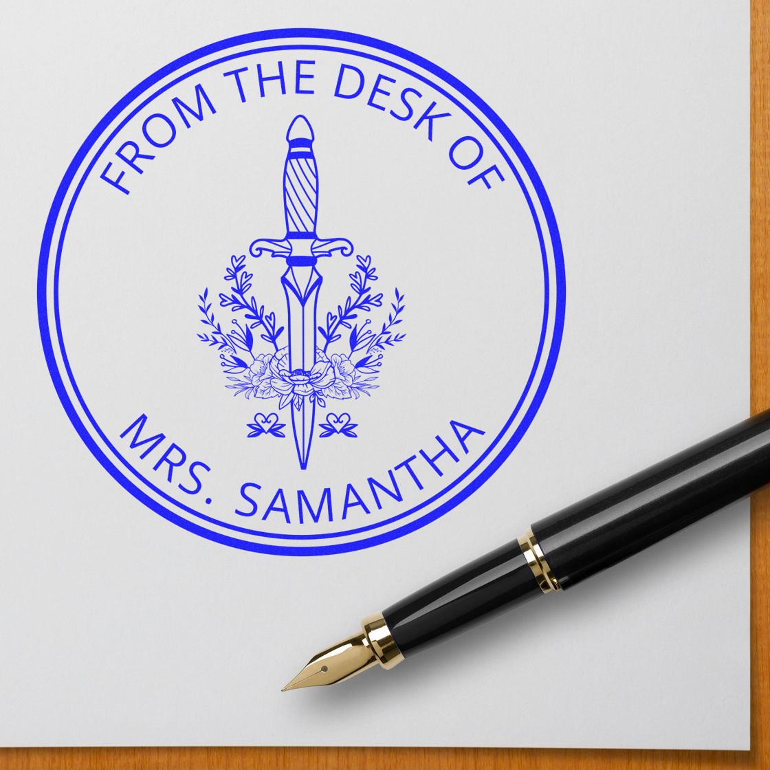 Blue custom desk stamp seal for Mrs. Samantha with sword and floral emblem, on white paper beside a black-and-gold fountain pen