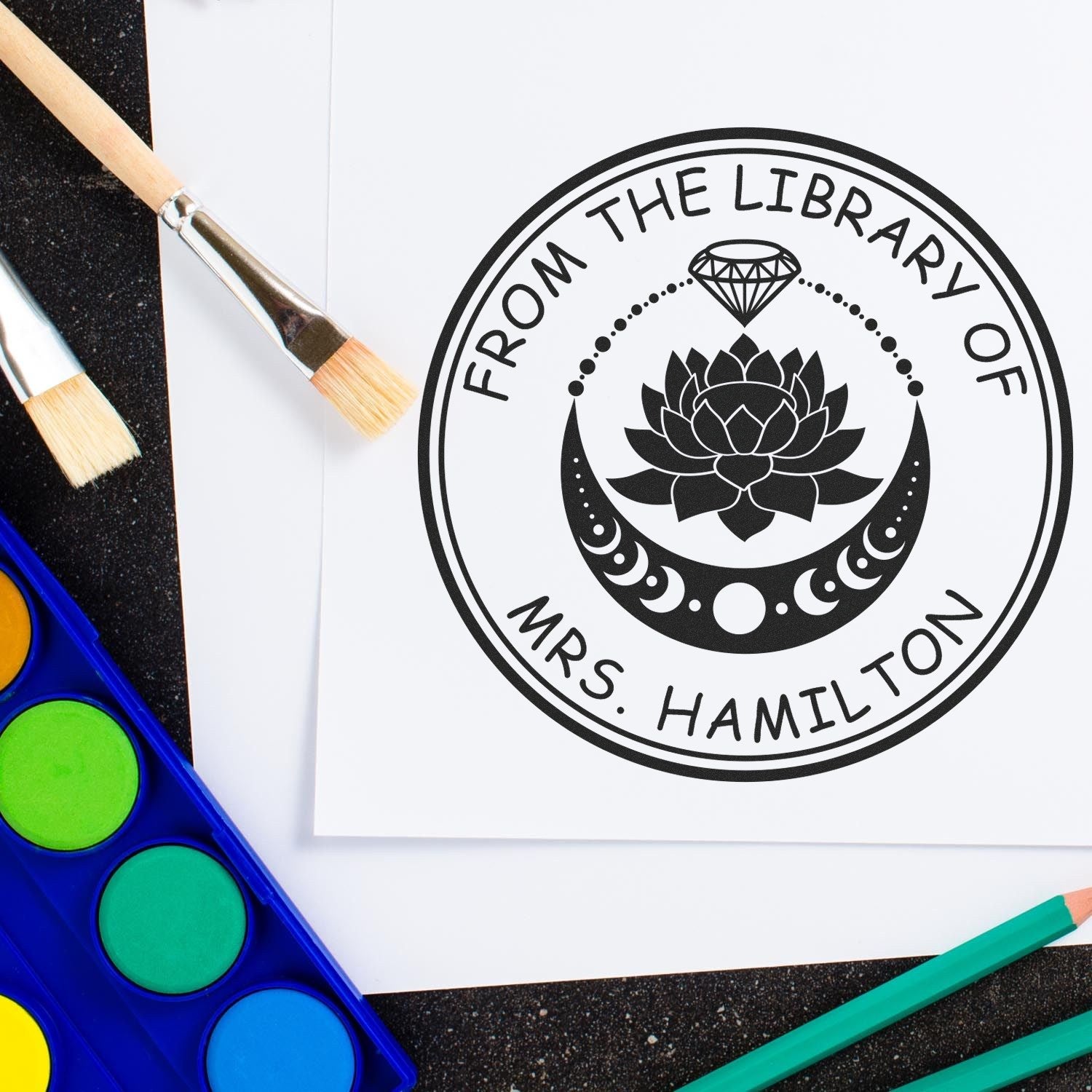 Black library stamp seal with lotus and diamond, reading From the Library of Mrs. Hamilton, beside brushes and watercolors.