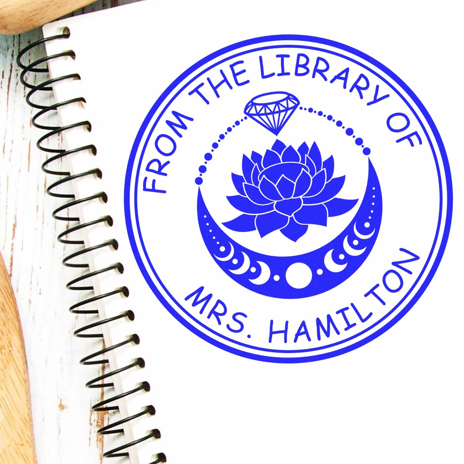 Blue circular library stamp seal reading From the Library of Mrs. Hamilton with lotus, gem, and moon motifs on white notebook.
