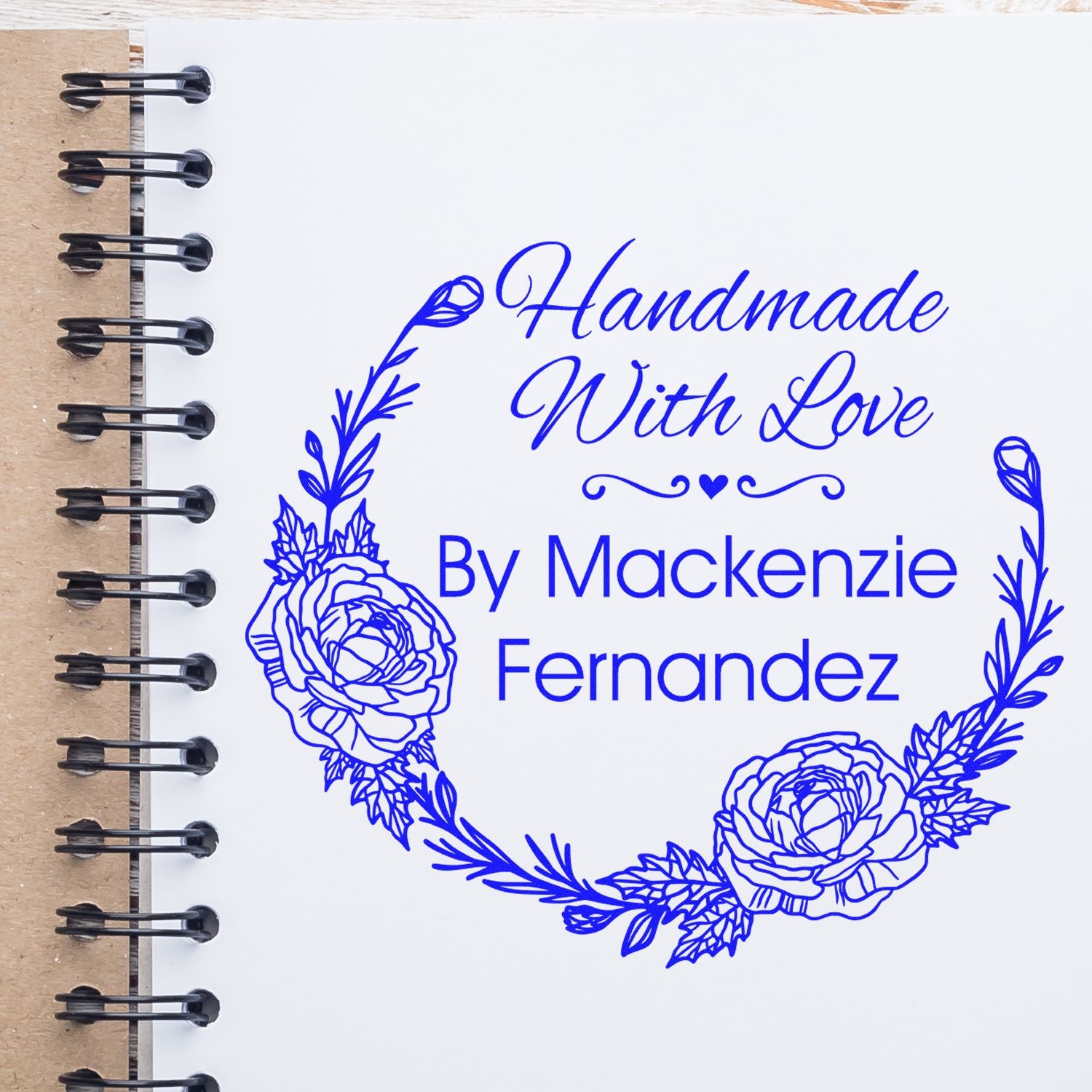 Blue handmade stamp seal with floral wreath and script text on white notebook page, reading Handmade With Love By Mackenzie Fernandez.