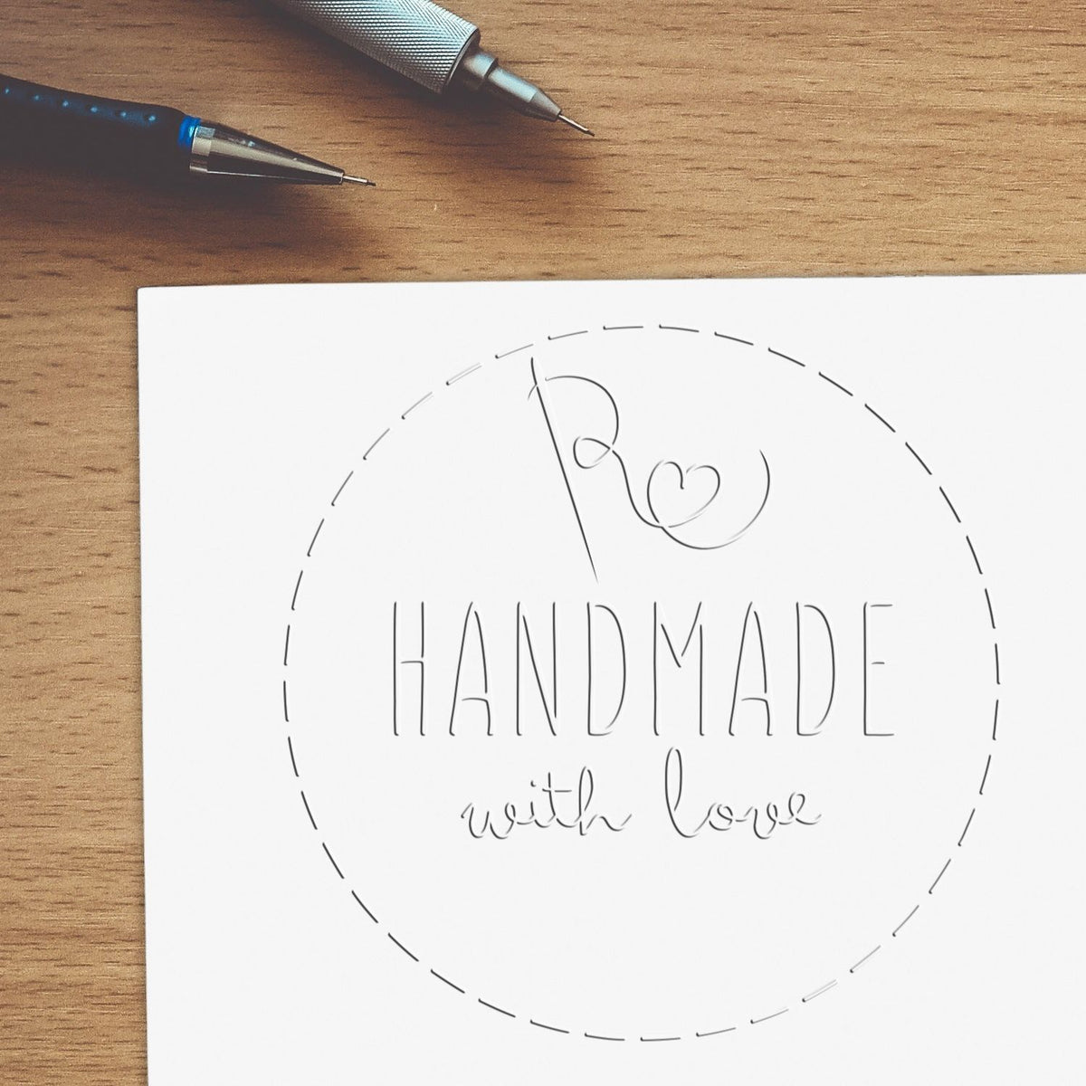 Custom stamp and seal design on white paper, reading HANDMADE with love, beside black and silver pens on a wood desk.
