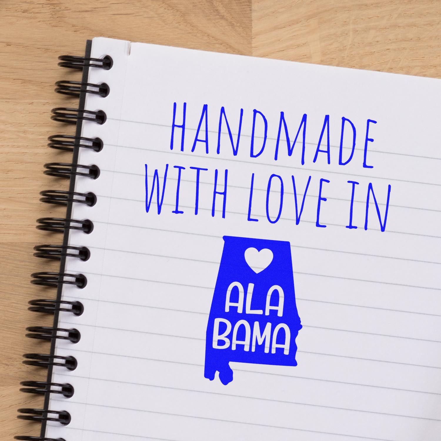 Wood Handle Alabama Handmade with Love Rubber Stamp on a notebook, featuring a blue design with the text Handmade with Love in Alabama and a heart symbol. Perfect for crafts and gifts.