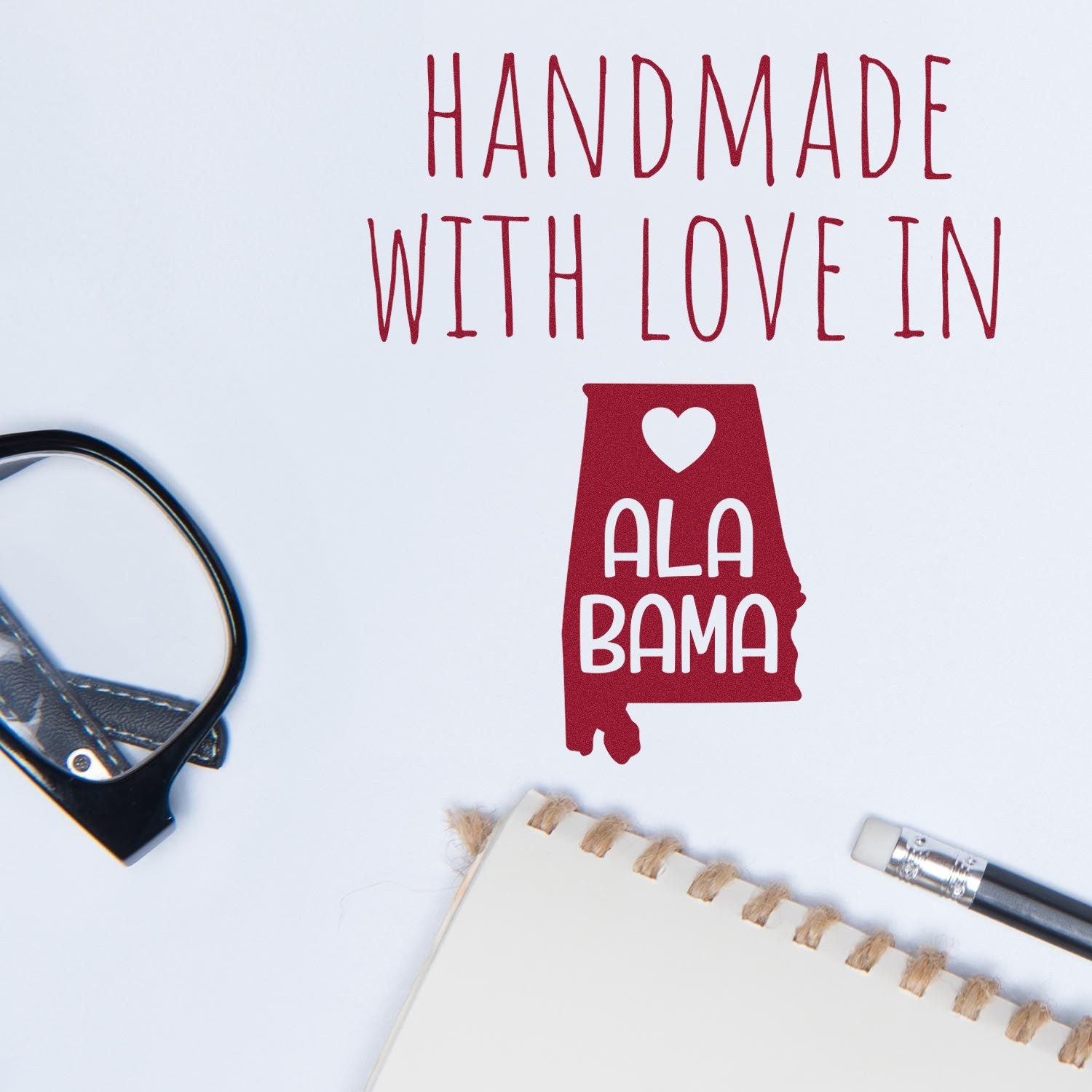 Wood Handle Alabama Handmade with Love Rubber Stamp on paper, featuring a red Alabama state outline with heart design, next to glasses and a notebook.