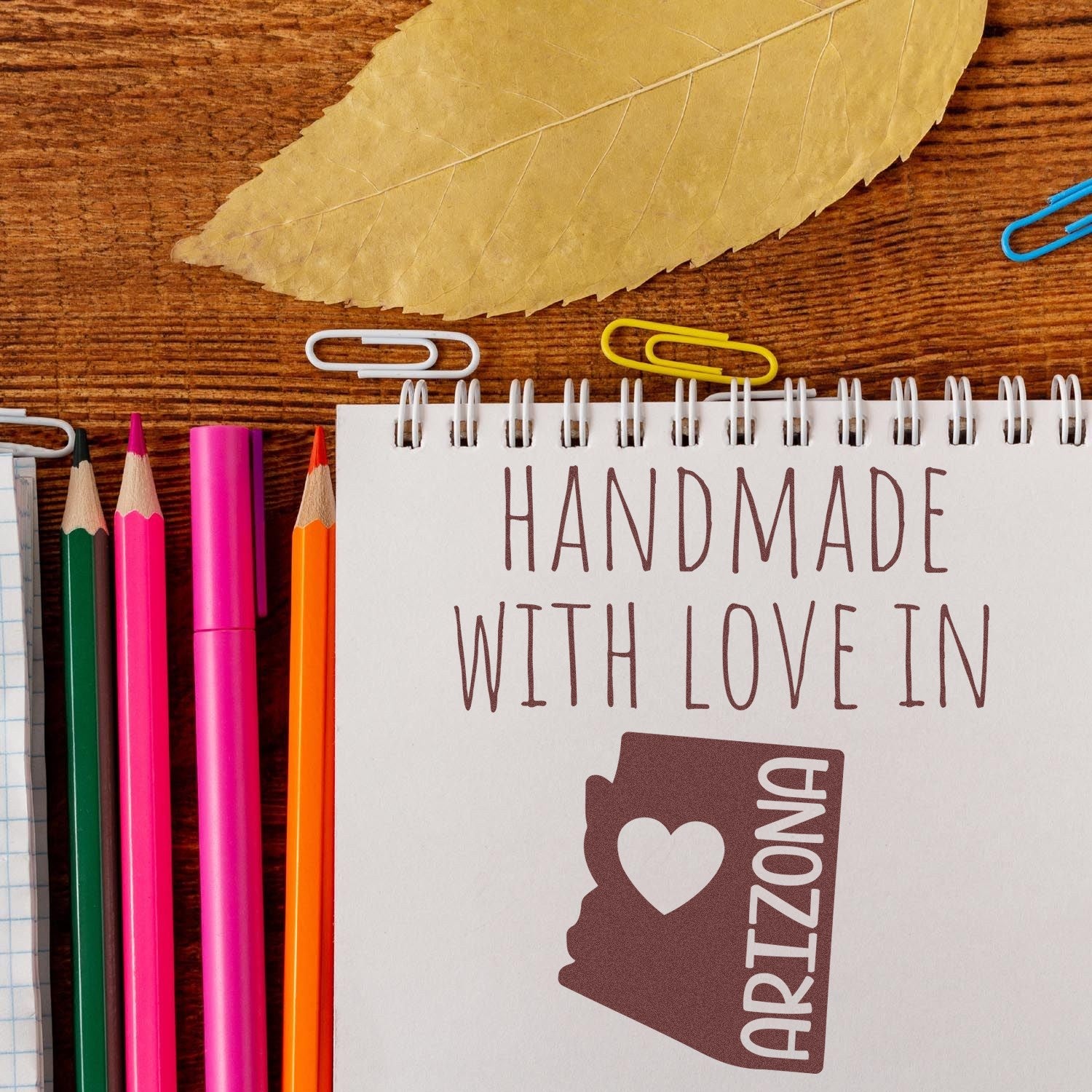 Self-Inking Arizona Handmade with Love Stamp on a notepad, surrounded by colorful pencils and paperclips, showcasing a heart and state outline design. Perfect for crafts and personalized gifts.