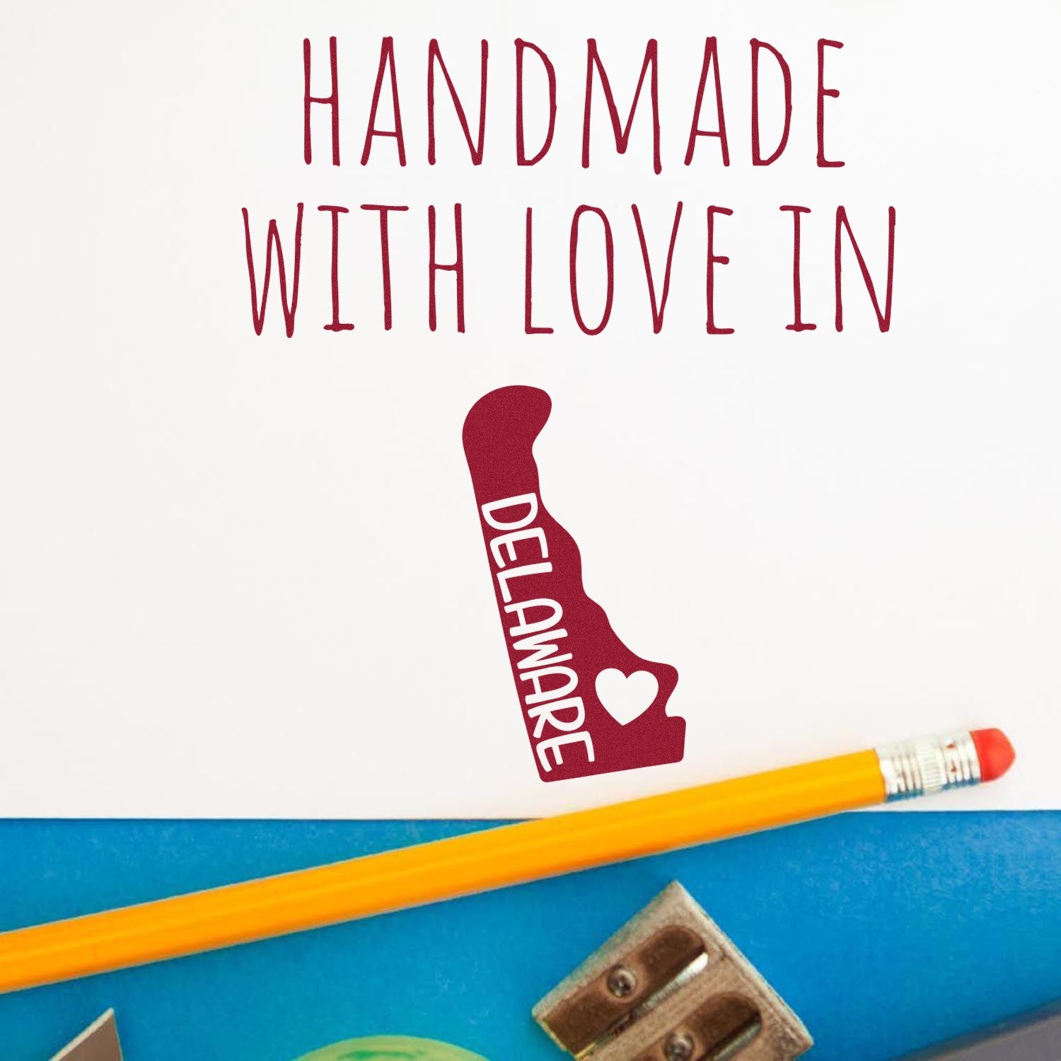 Image of a PSI Pre-Inked stamp with Handmade with Love in Delaware text, featuring a Delaware state outline and heart, alongside a pencil and sharpener on a blue surface.