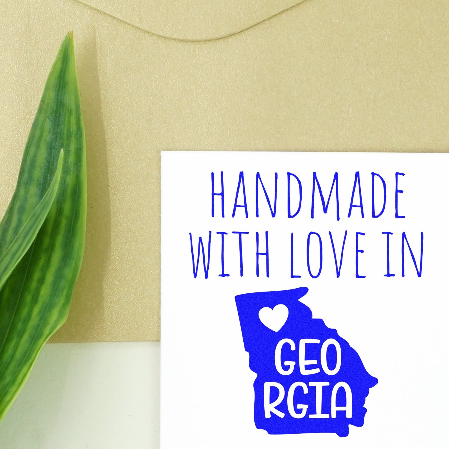 Handmade with Love in Georgia Slim Pre-Inked Stamp featuring a blue Georgia state outline with a heart, placed on a beige background with a green leaf. Perfect for personalized crafts.