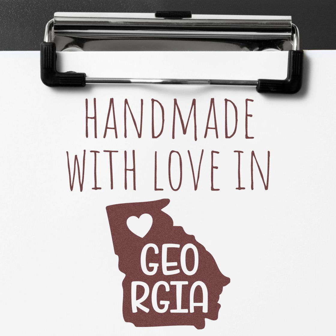 Self-Inking Georgia Handmade with Love Stamp featuring a heart and Georgia outline, perfect for crafts and gifts. Brown ink on white paper with a clipboard.