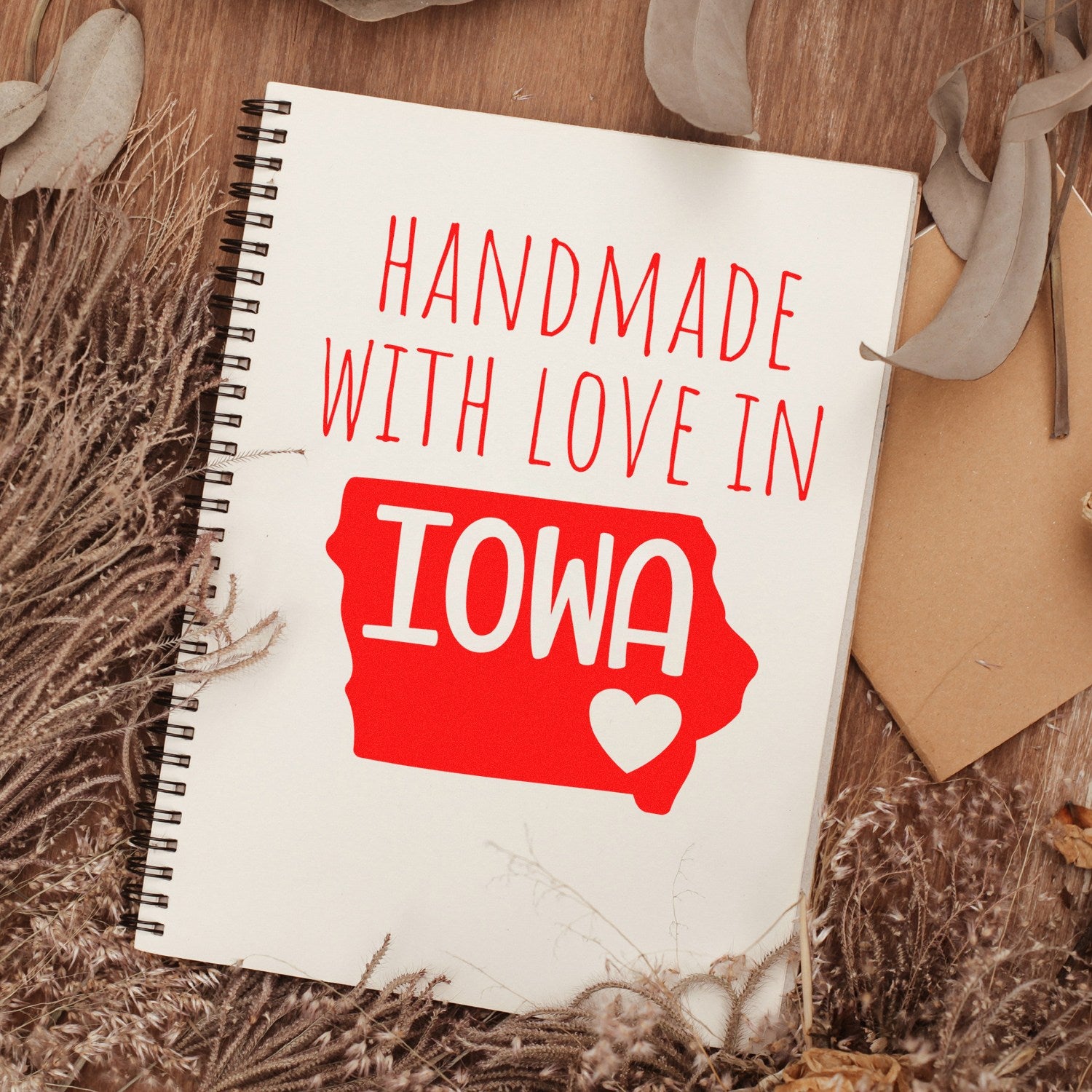 Handmade with Love in Iowa Slim Pre-Inked Stamp on a notebook, featuring red text and a heart on a map of Iowa, surrounded by dried flowers and a kraft envelope.