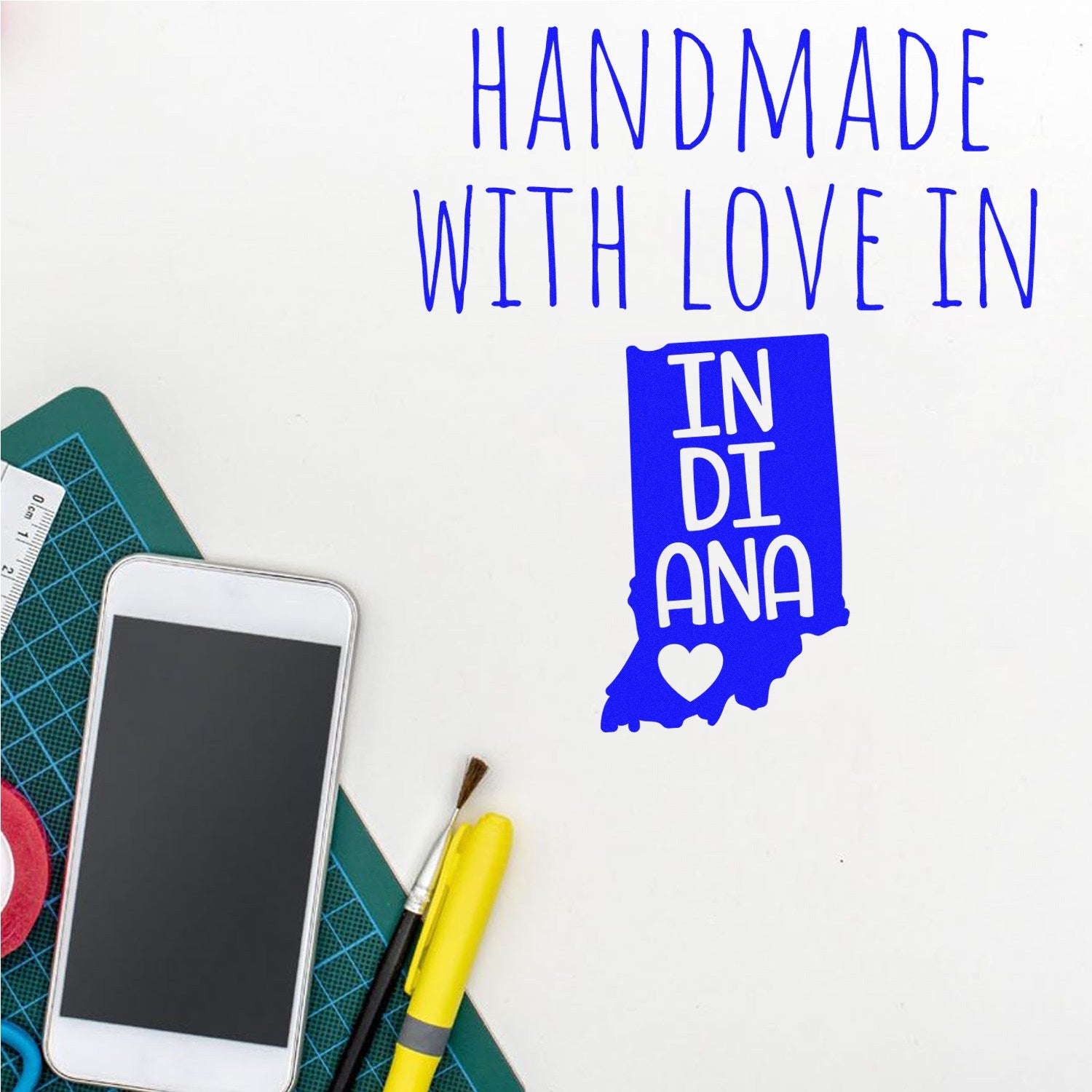 PSI Pre-Inked Handmade with Love in Indiana stamp, featuring a blue Indiana state outline with a heart, next to a smartphone and crafting tools on a desk.