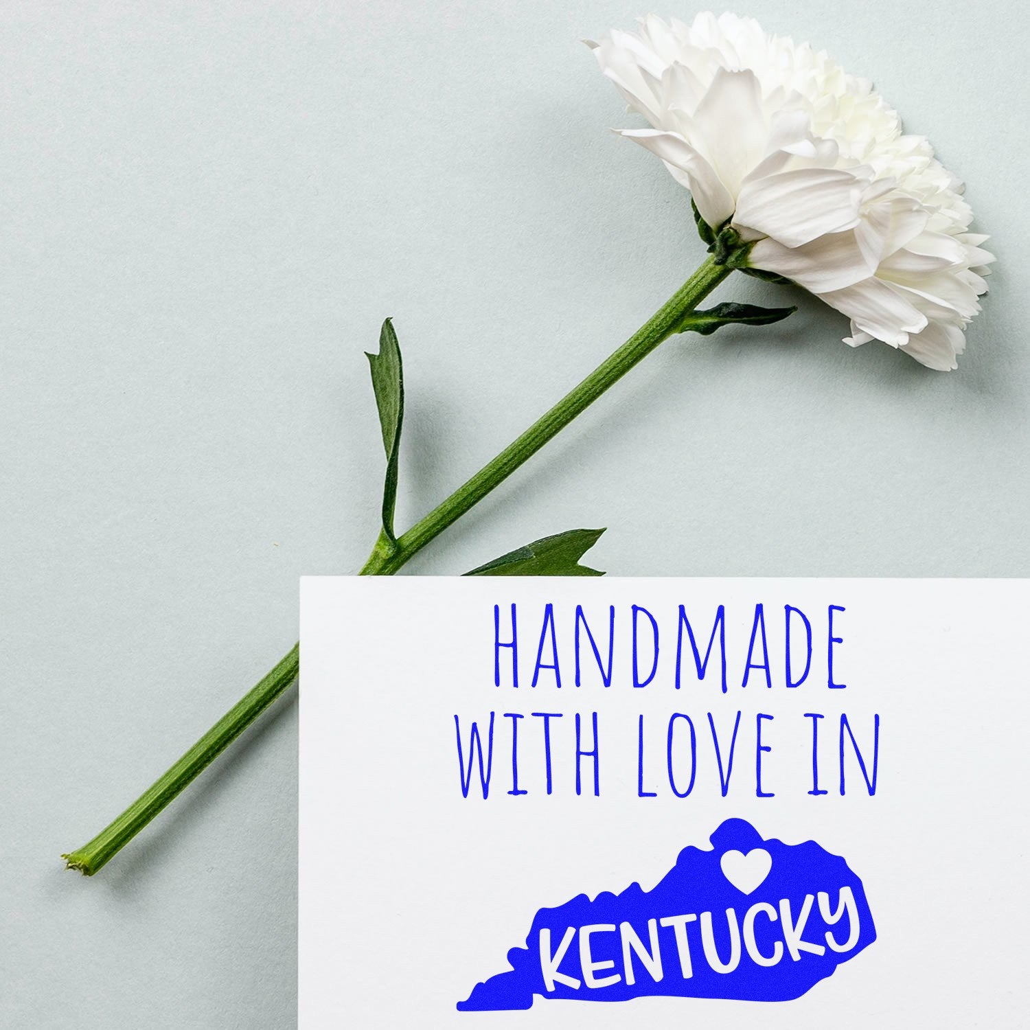 Wood Handle Kentucky Handmade with Love Rubber Stamp on paper with blue text and Kentucky outline, next to a white flower on a light background.