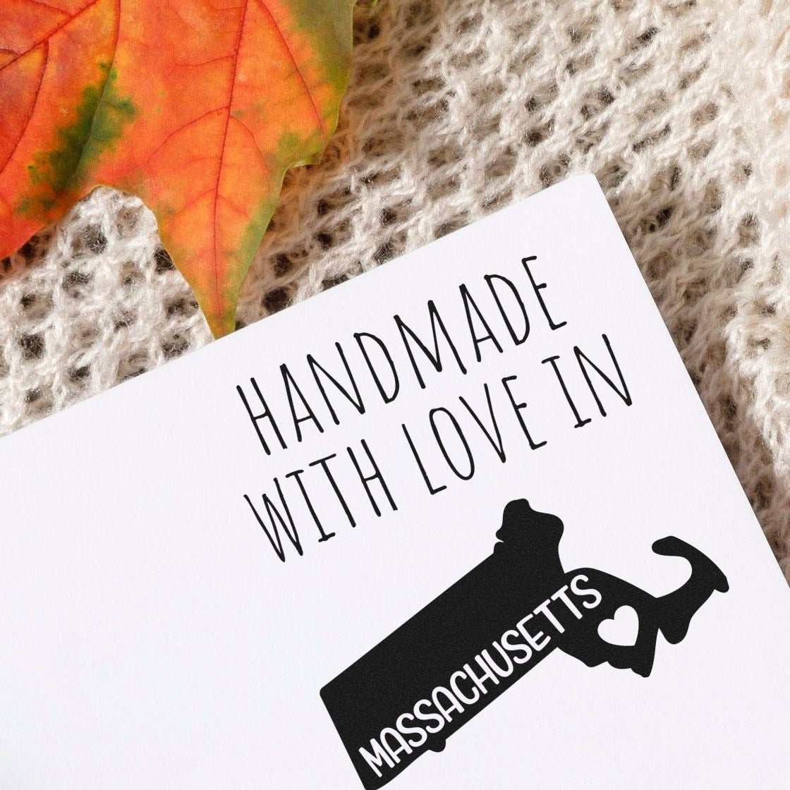 PSI Pre-Inked Handmade with Love in Massachusetts stamp on white card, featuring a black silhouette of Massachusetts with a heart, placed on a knitted fabric with a fall leaf.