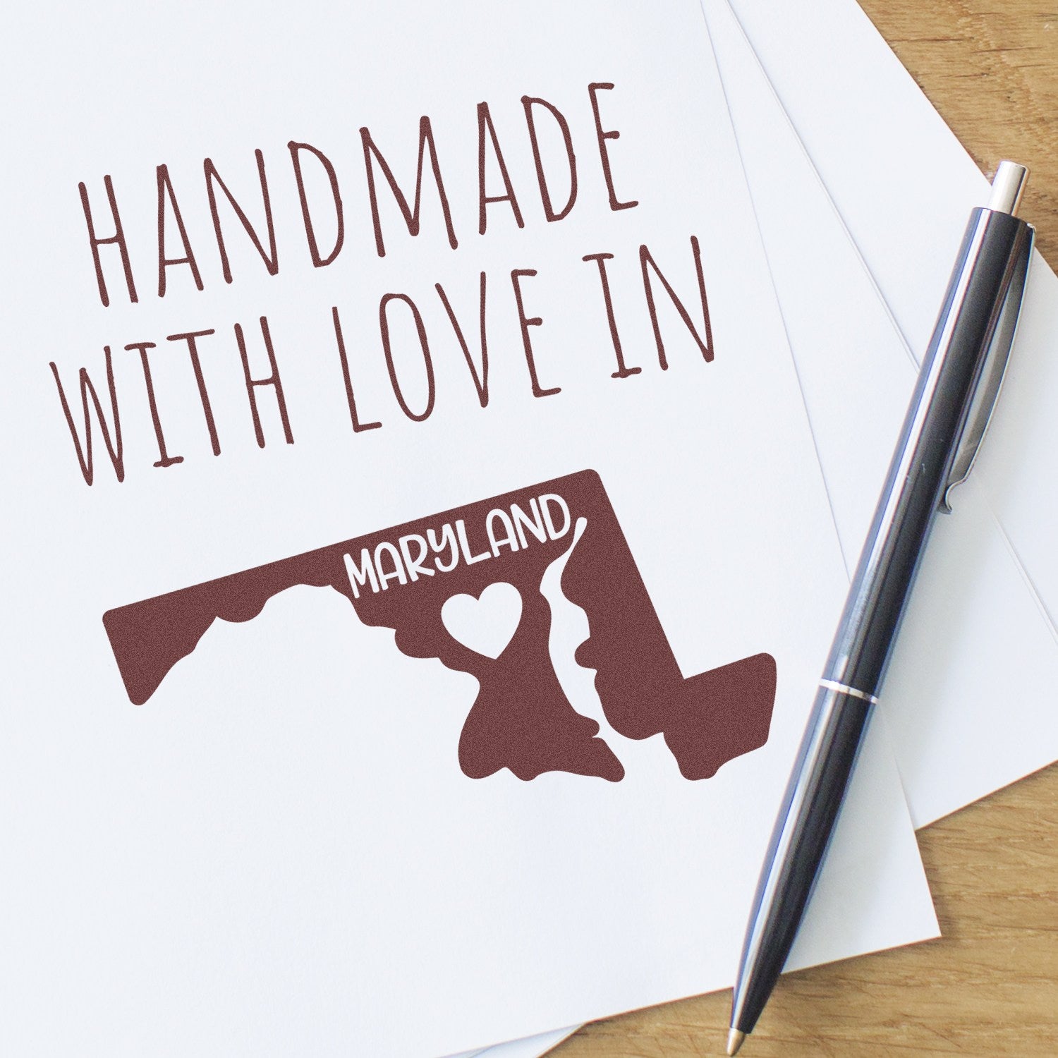 Self-Inking Maryland Handmade with Love Stamp featuring a Maryland map design with heart, shown on white paper with a pen. Perfect for crafts and personalized projects.