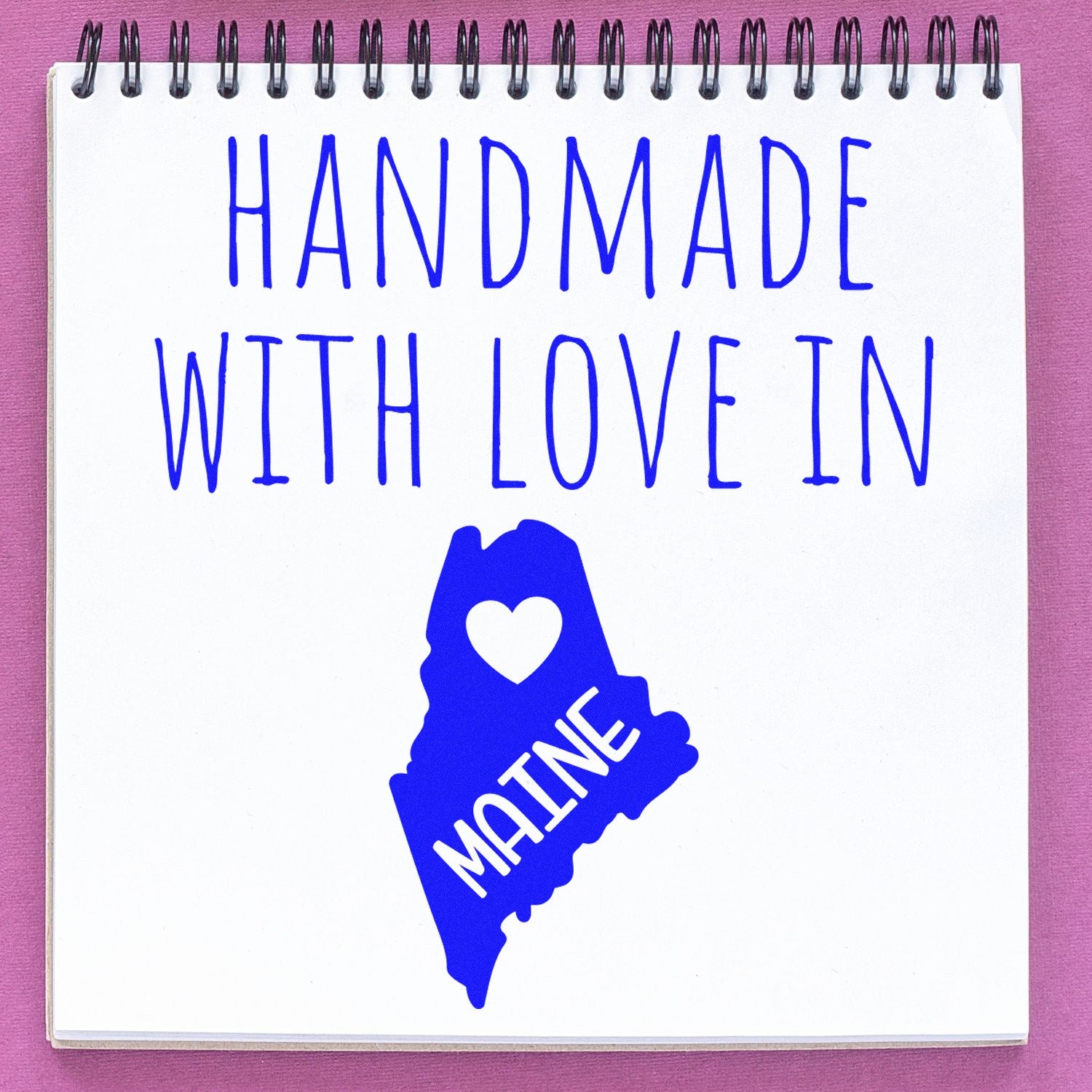 Wood Handle Maine Handmade with Love Rubber Stamp on a notepad, featuring a blue design with Handmade with Love in Maine and a heart inside the state outline. Perfect for crafts and gifts.