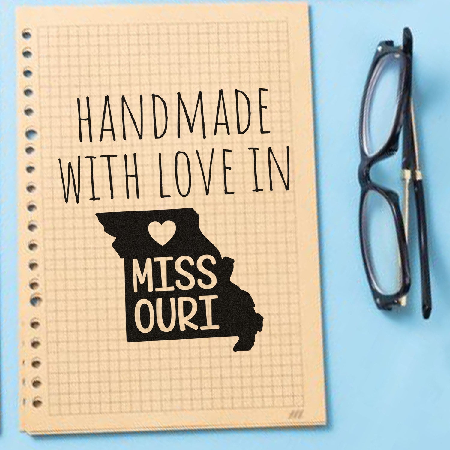 Self-Inking Missouri Handmade with Love Stamp on grid paper, featuring a heart and Missouri outline, next to black glasses on a blue background.