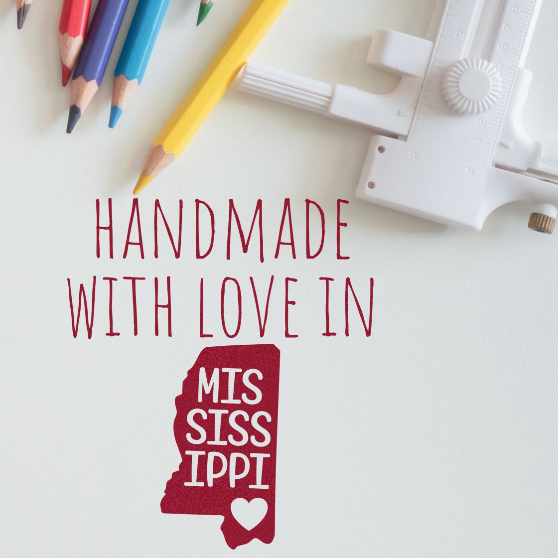 Handmade with Love in Mississippi Slim Pre-Inked Stamp featuring a red Mississippi state outline with heart, surrounded by colorful pencils and a ruler on a white background.