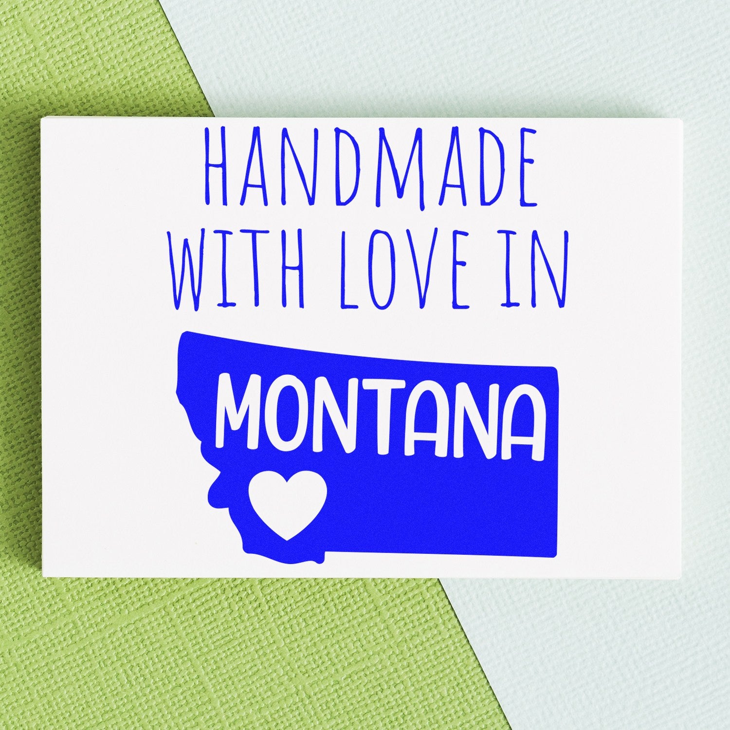 Handmade with Love in Montana Slim Pre-Inked Stamp on a textured background, featuring a blue Montana state outline with a heart. Perfect for crafts and personalized projects.