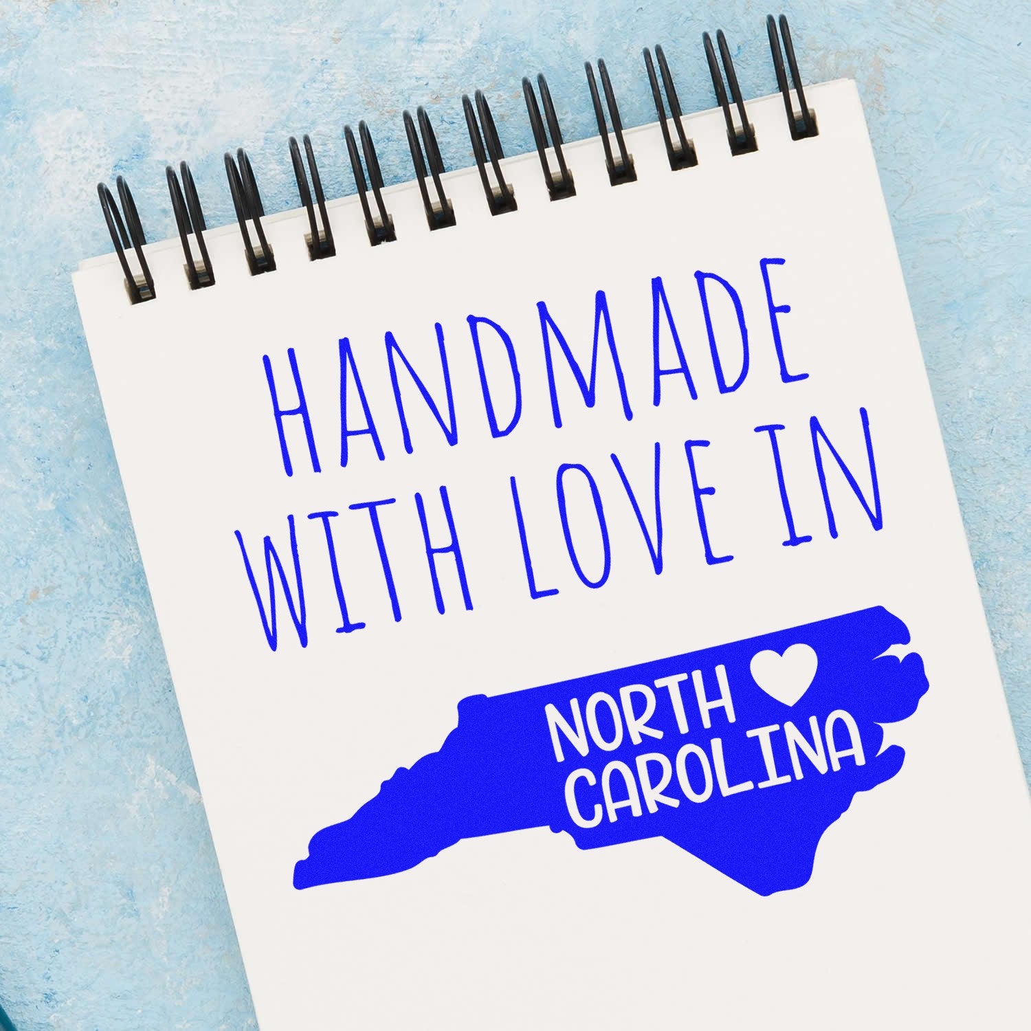 Wood Handle North Carolina Handmade with Love Rubber Stamp on a notepad, featuring a blue map of North Carolina with a heart. Perfect for crafts and personalized projects.