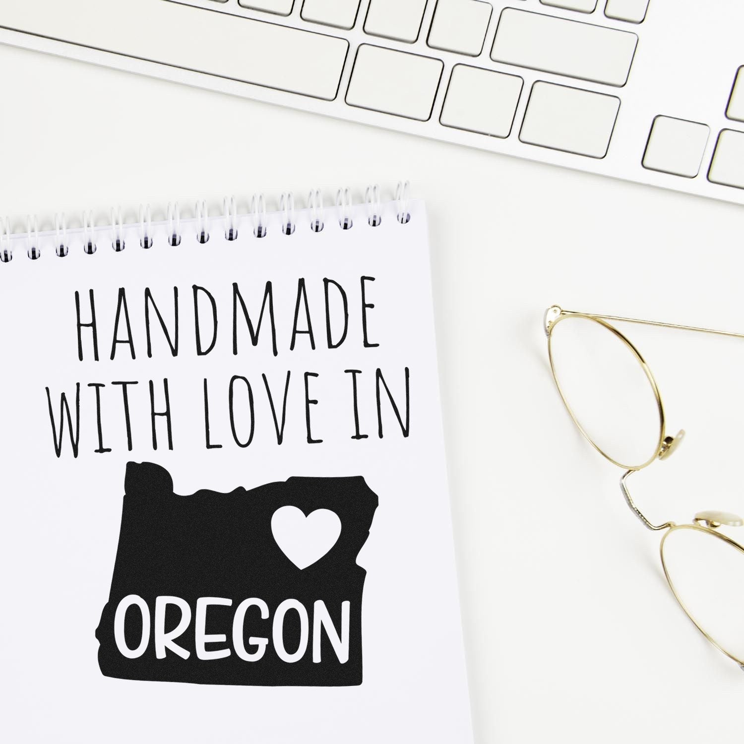 Self-Inking Oregon Handmade with Love Stamp on a notepad, featuring a heart and Oregon state outline. Perfect for crafts and gifts. Keyboard and glasses in the background.