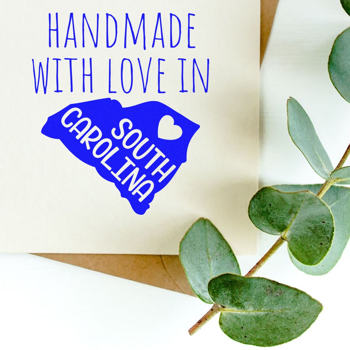 Self-Inking South Carolina Handmade with Love Stamp on paper, featuring blue text and heart design, surrounded by green leaves.