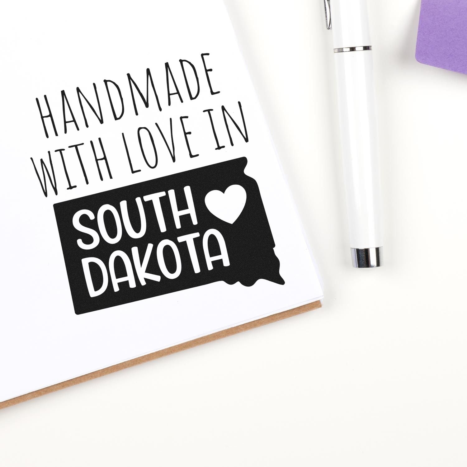 Wood Handle South Dakota Handmade with Love Rubber Stamp on paper, featuring a heart and state outline, next to a pen and purple notepad. Perfect for crafts and personalized gifts.