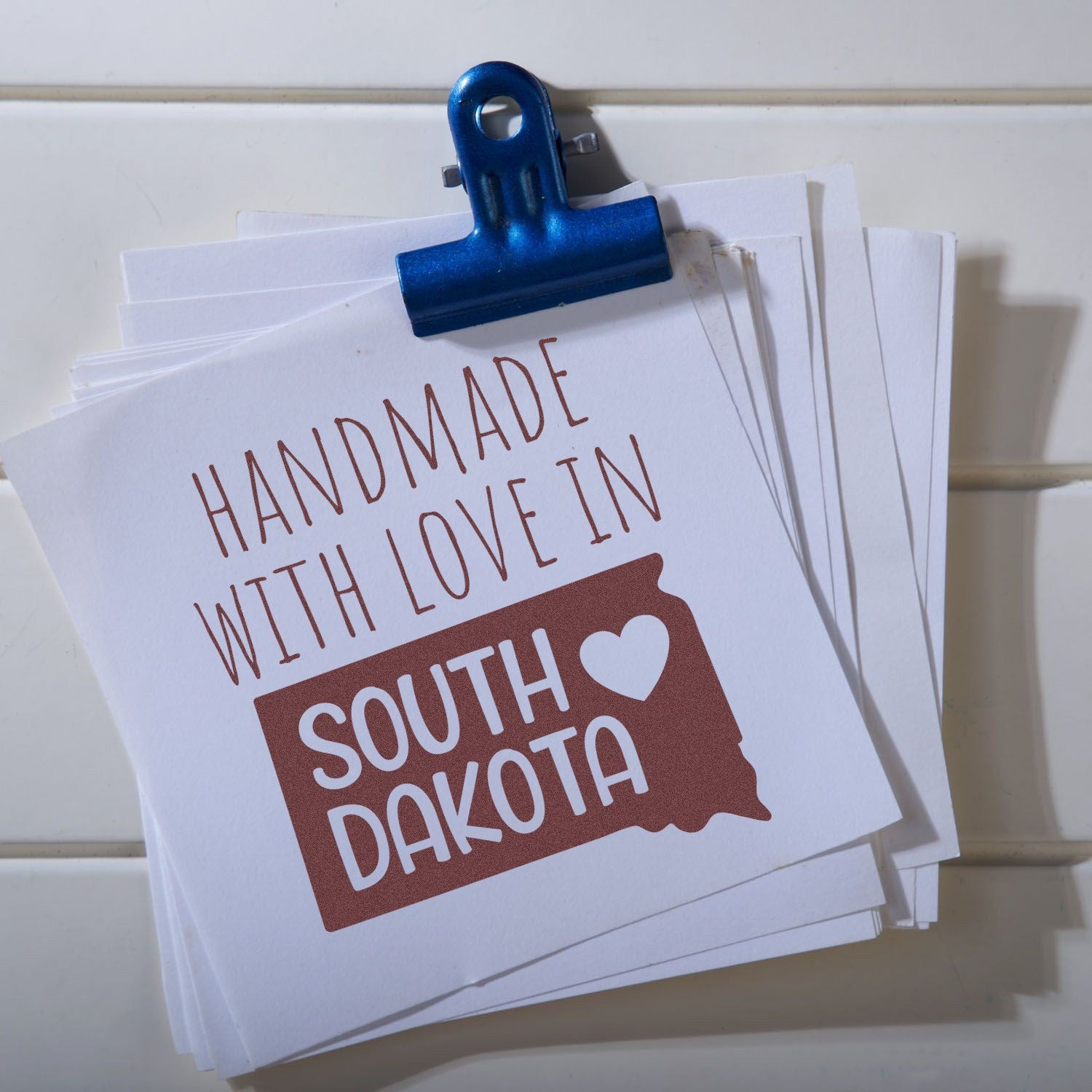 Wood Handle South Dakota Handmade with Love Rubber Stamp on white paper, featuring a heart and state outline, clipped with a blue holder.