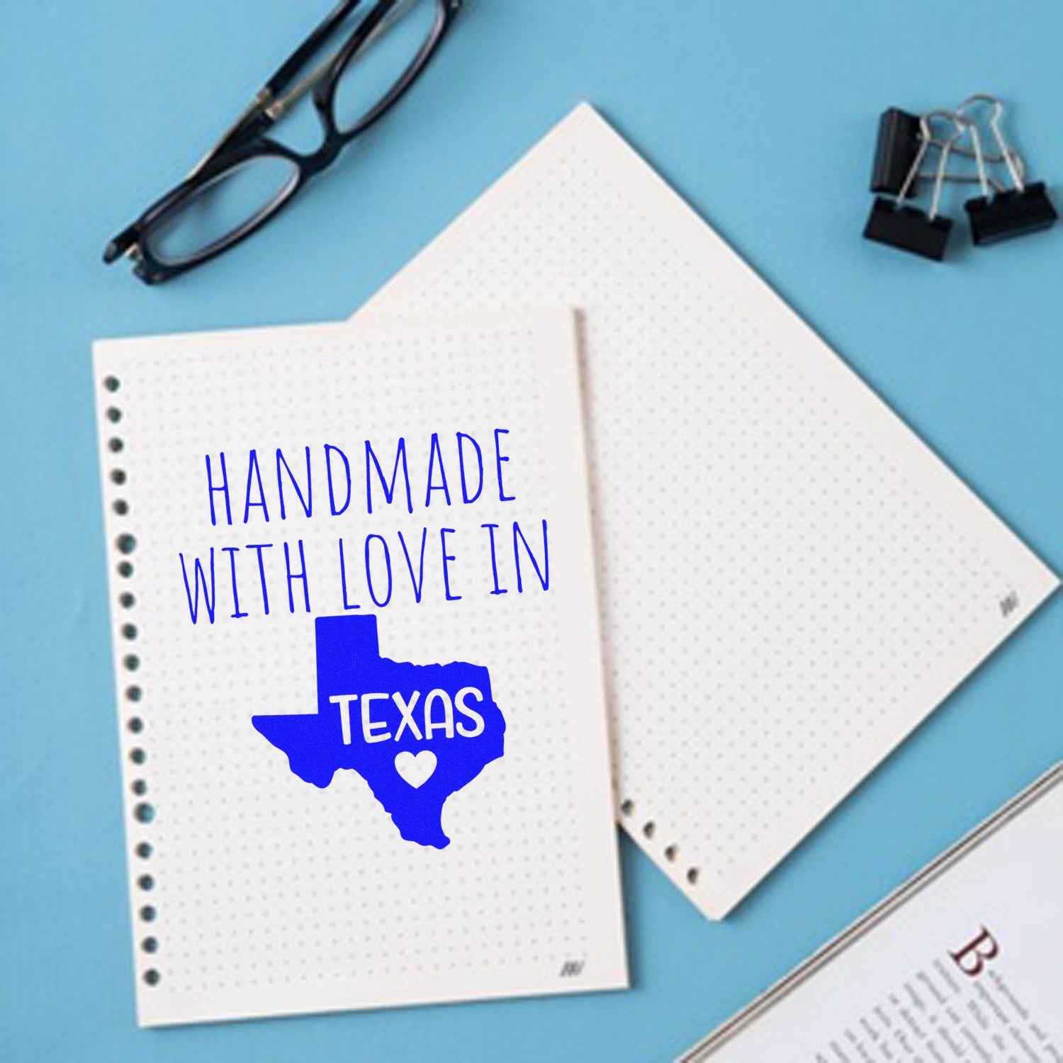 Handmade with Love in Texas Slim Pre-Inked Stamp on a blue background, next to glasses and notepads, showcasing its design and craftsmanship.