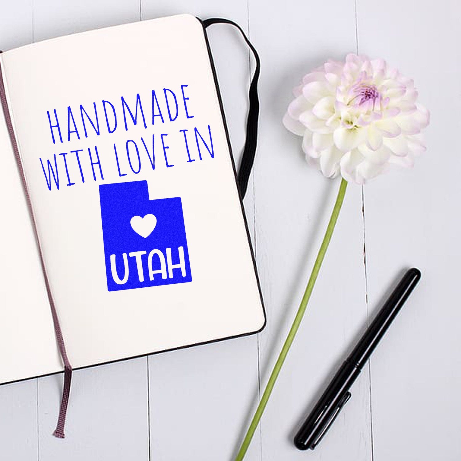 Handmade with Love in Utah Slim Pre-Inked Stamp on open notebook, featuring blue state outline and heart design, next to a white flower on a light wooden surface.