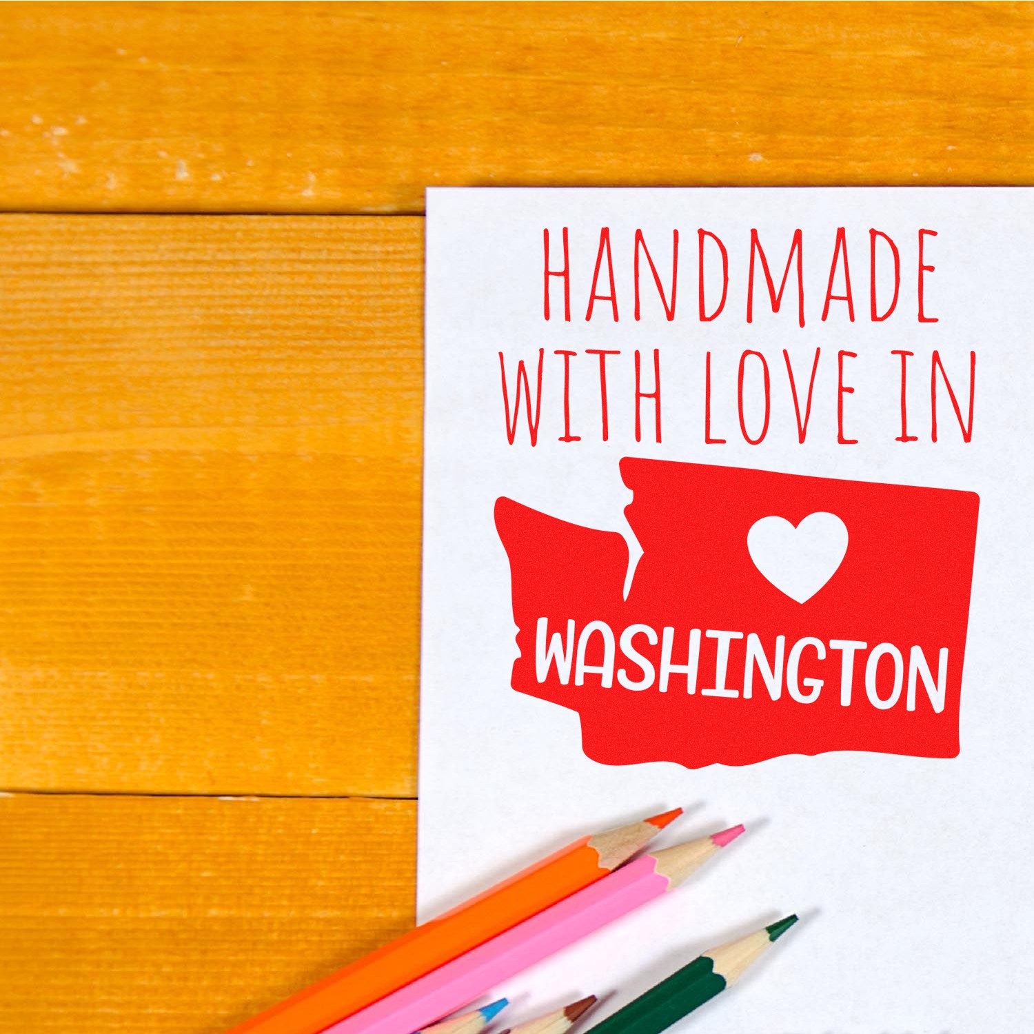 Self-Inking Washington Handmade with Love Stamp on paper, featuring a red Washington state outline with a heart and text. Includes colored pencils on a wooden surface.