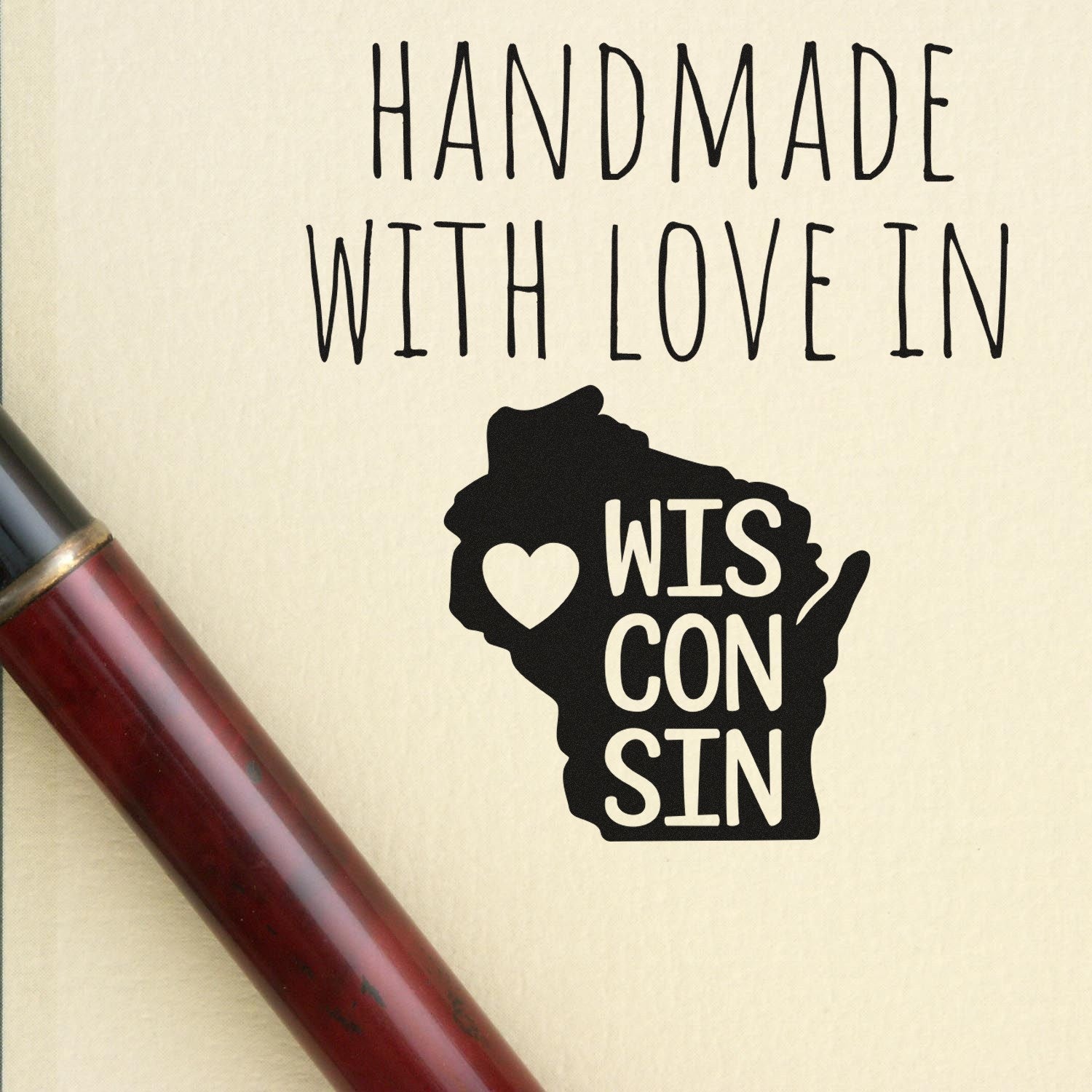 Self-Inking Wisconsin Handmade with Love Stamp featuring a heart inside the state outline, perfect for crafts. Includes a sleek black design on a beige background with a pen beside it.