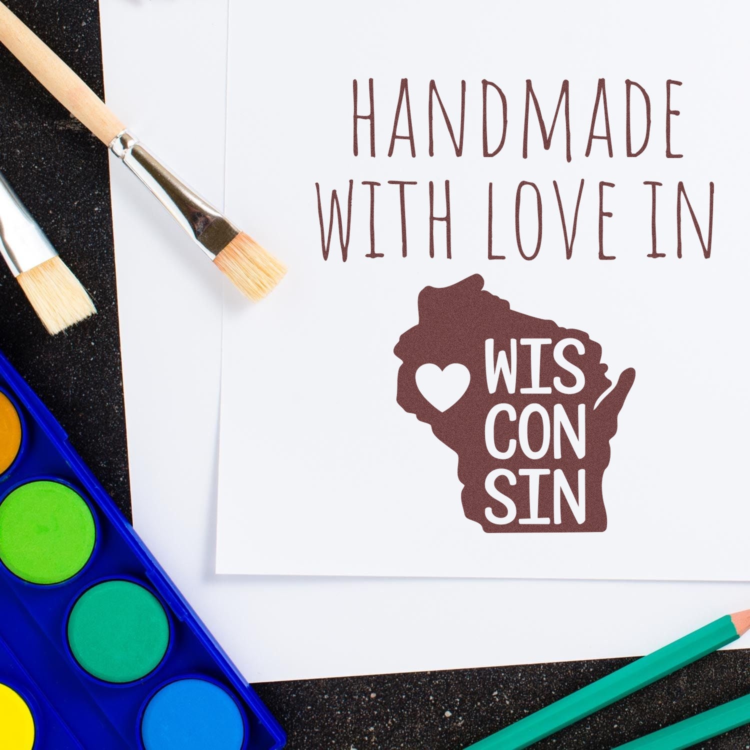 Self-Inking Wisconsin Handmade with Love Stamp featuring a heart and state outline, surrounded by art supplies on a black surface.