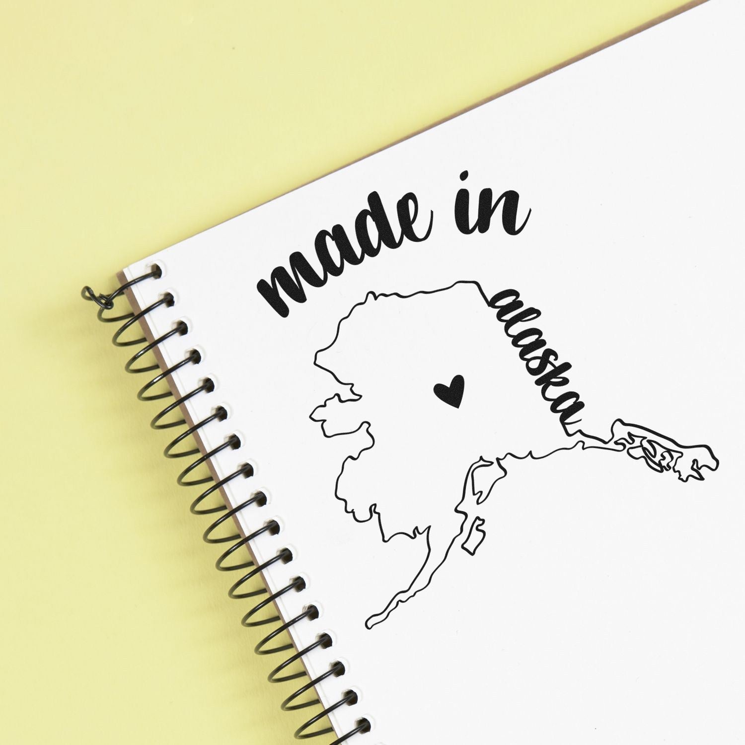 Slim Pre-Inked Stamp Alaska Made in Stamp on a notebook, featuring a heart and Alaska outline. Perfect for adding a personal touch to stationery.