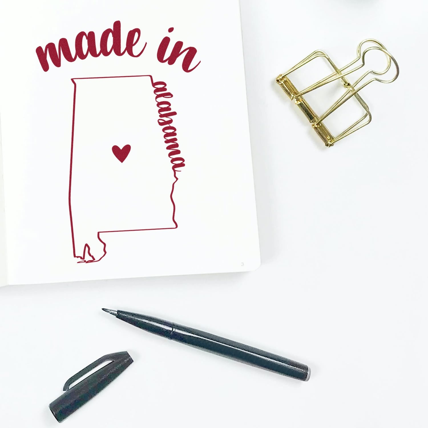 Made with Love in Alabama Rubber Stamp featuring a heart inside the Alabama state outline, perfect for crafts and gifts. Includes a pen and binder clip for scale.