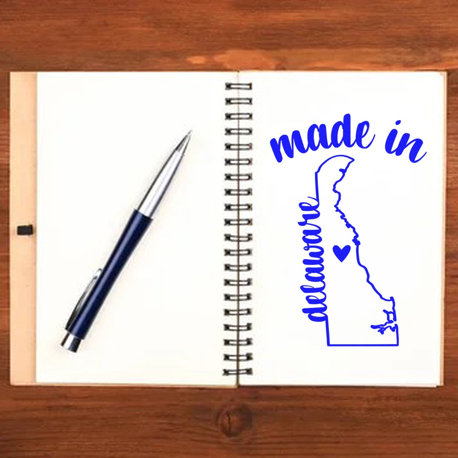 Made with Love in Delaware Rubber Stamp on a notebook, featuring a blue outline of Delaware with a heart, and a pen beside it on a wooden surface.