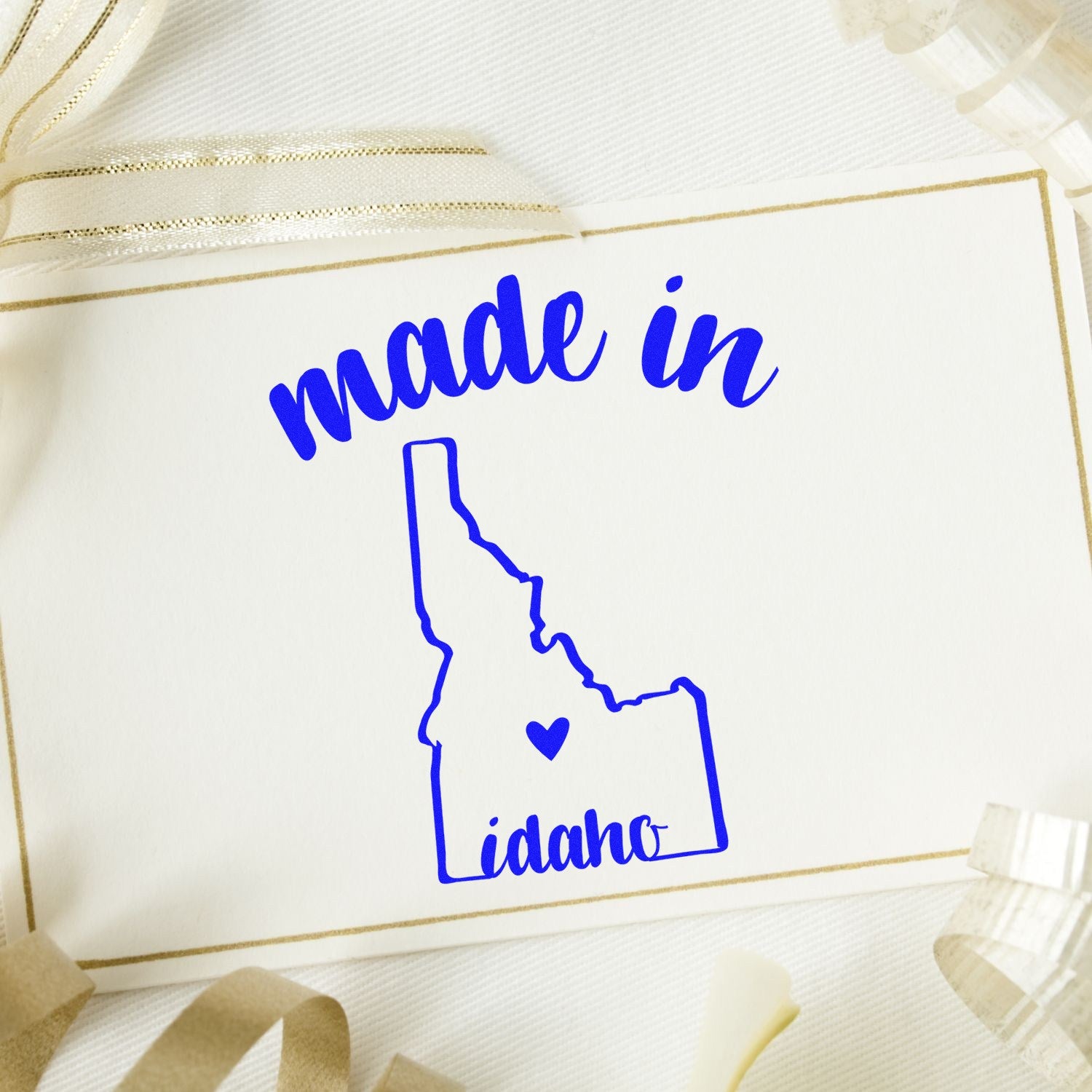 Made in Idaho Stamp Pre-Inked: Blue stamp with 'made in' text and Idaho state outline on a card, surrounded by ribbon. Perfect for crafts and gifts.