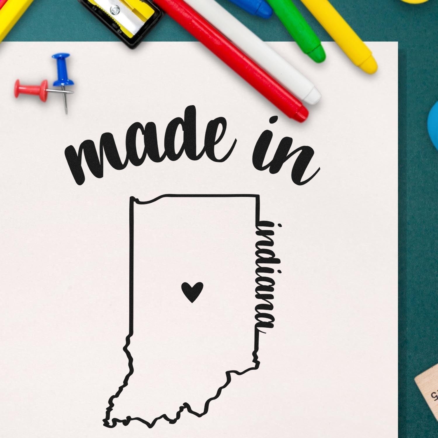 Made with Love in Indiana Rubber Stamp featuring Indiana state outline with heart, surrounded by colorful pens and pushpins on a green surface.