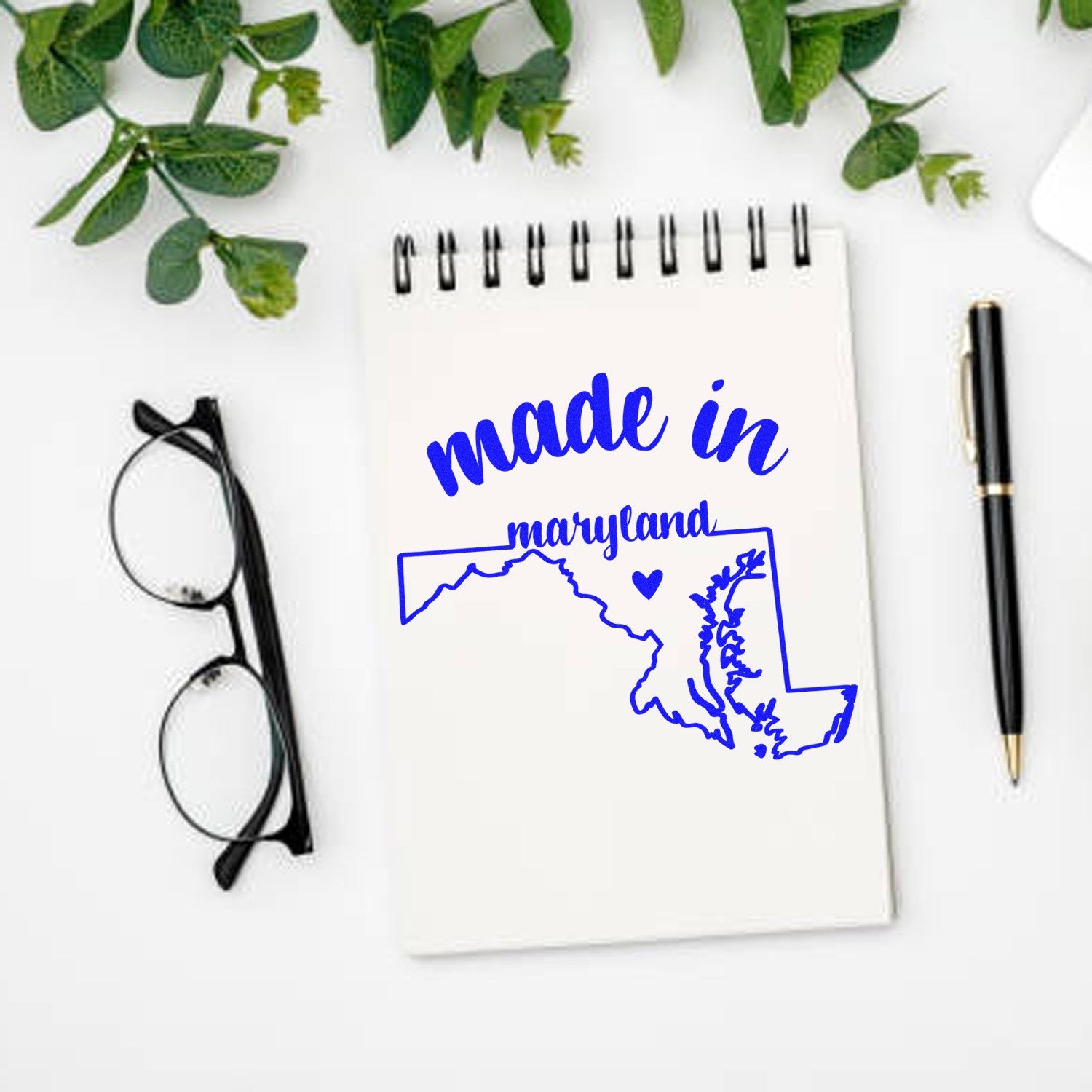 Made in Maryland Stamp Pre-Inked, featuring a blue outline of Maryland with 'made in' text, displayed on a notepad with glasses and a pen nearby.