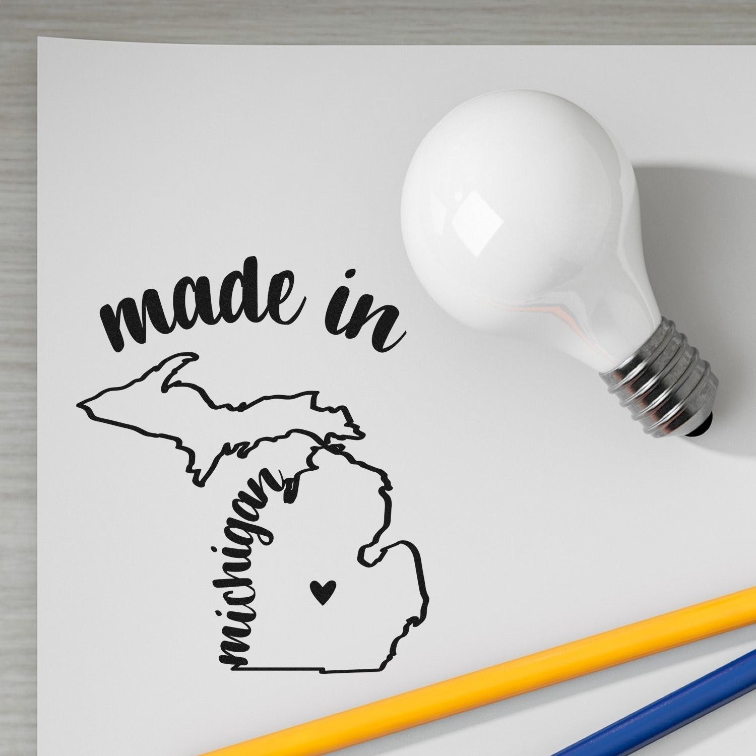 Made with Love in Michigan Rubber Stamp featuring a map outline of Michigan with 'made in' text, displayed on paper beside a light bulb and pencils.