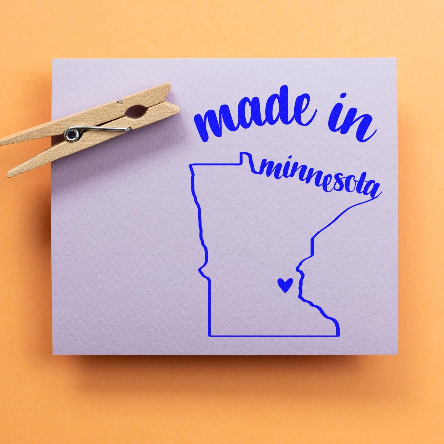 Made with Love in Minnesota Rubber Stamp featuring a blue outline of Minnesota with a heart, and the words 'made in minnesota' on a light purple background with a clothespin.