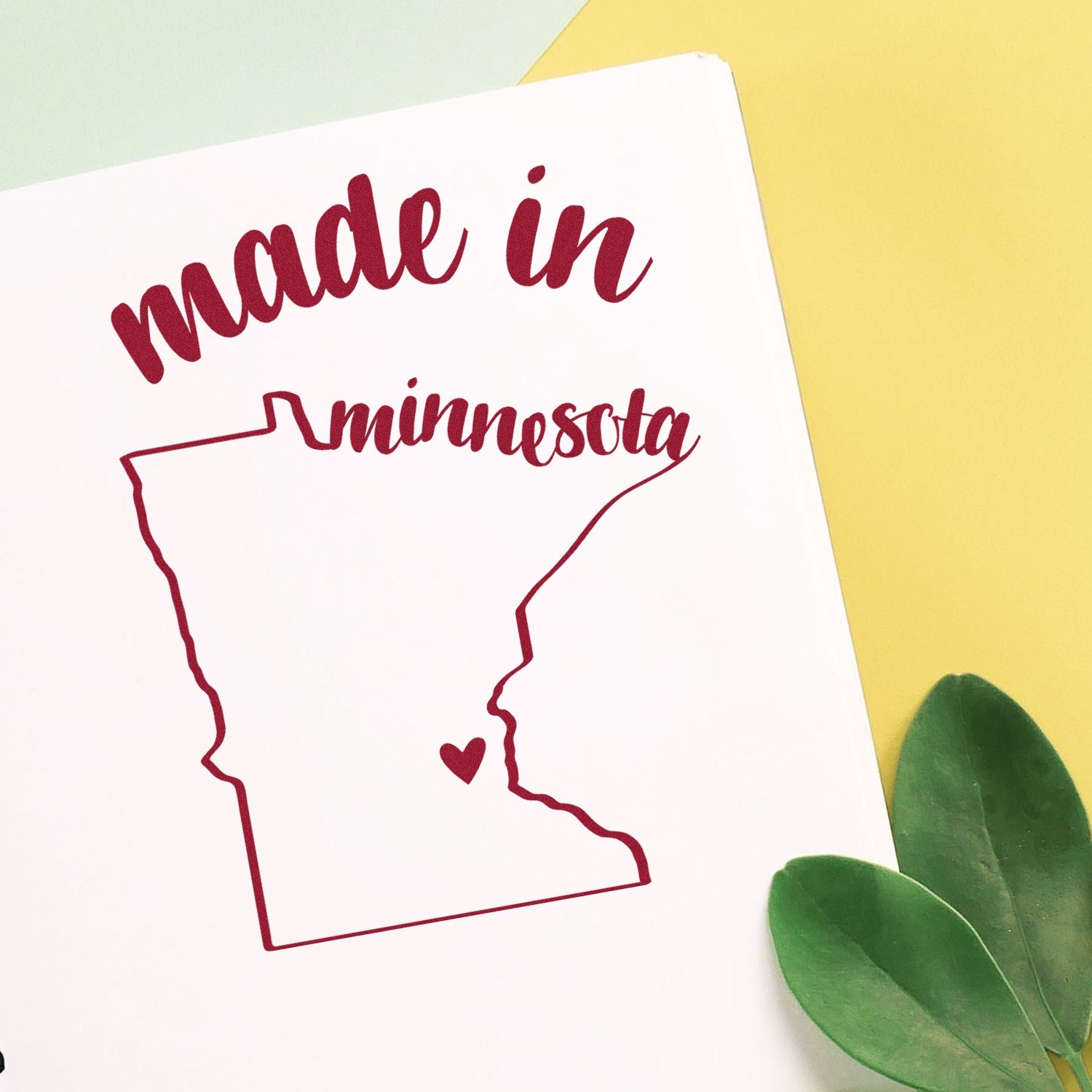 Made with Love in Minnesota Rubber Stamp featuring a red outline of Minnesota with a heart, set against a white background with green leaves and yellow paper accents.