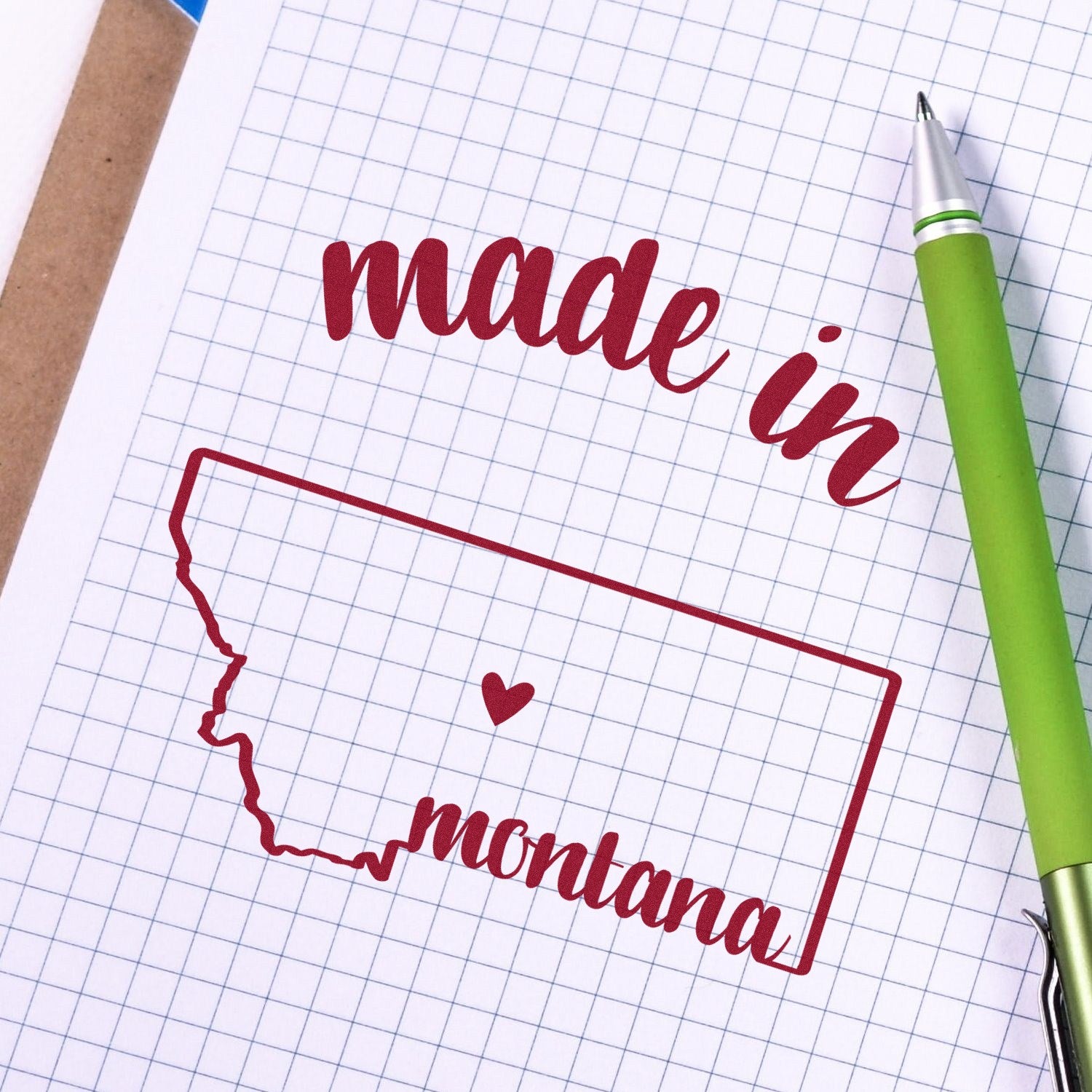 Slim Pre-Inked Stamp Montana Made in Stamp on graph paper, featuring a red outline of Montana with made in montana text and a heart, next to a green pen.