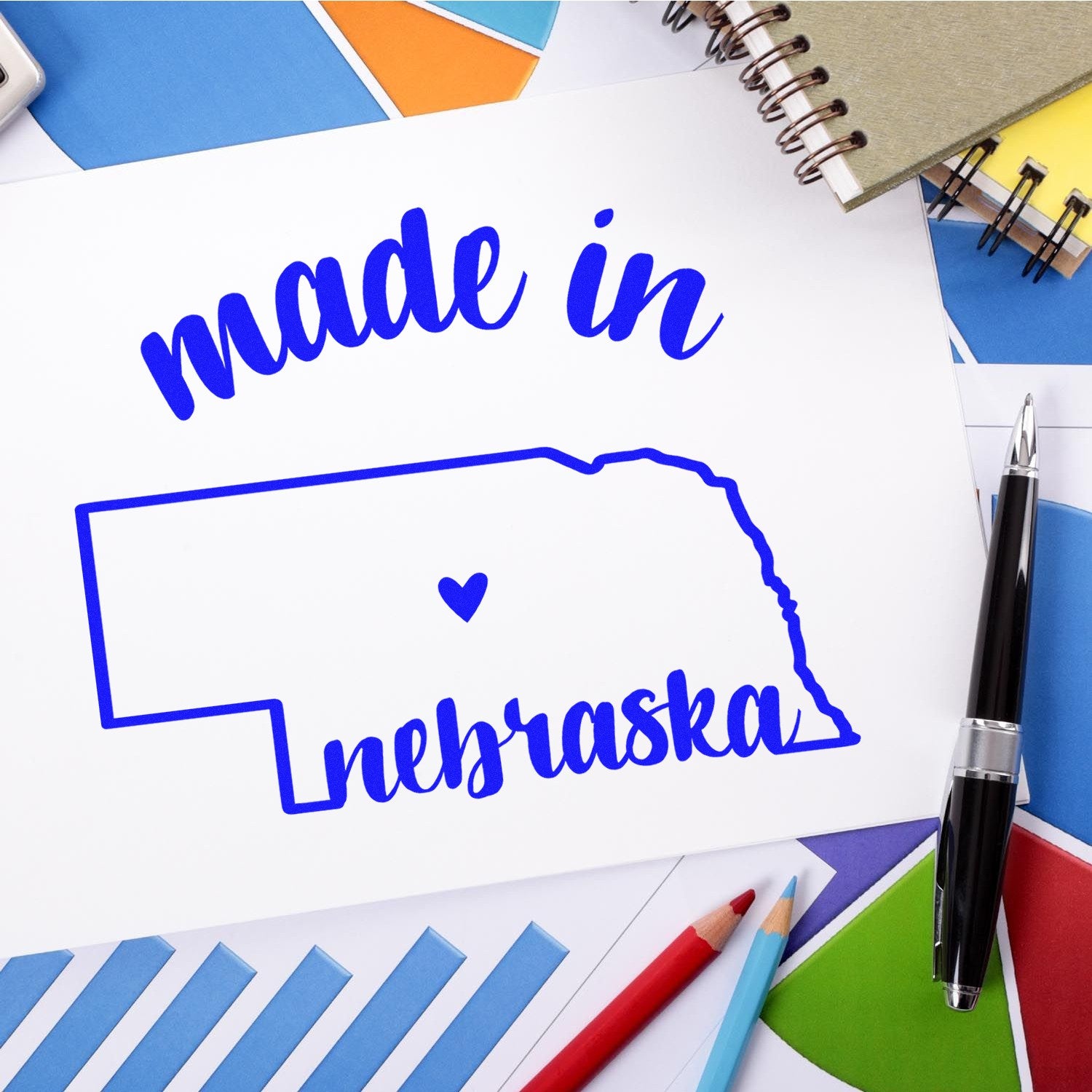 Made in Nebraska Stamp Pre-Inked featuring a blue outline of Nebraska with 'made in' and 'nebraska' text, displayed on a desk with colorful papers and a pen.