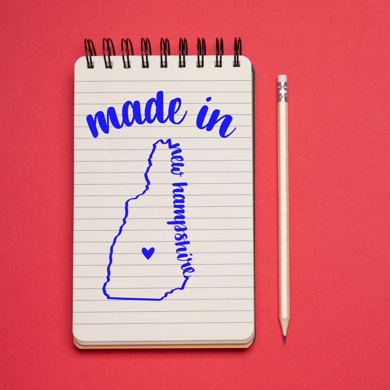 Image of the Made with Love in New Hampshire Rubber Stamp featuring a blue outline of New Hampshire with a heart, stamped on a notepad with a pencil on a red background.