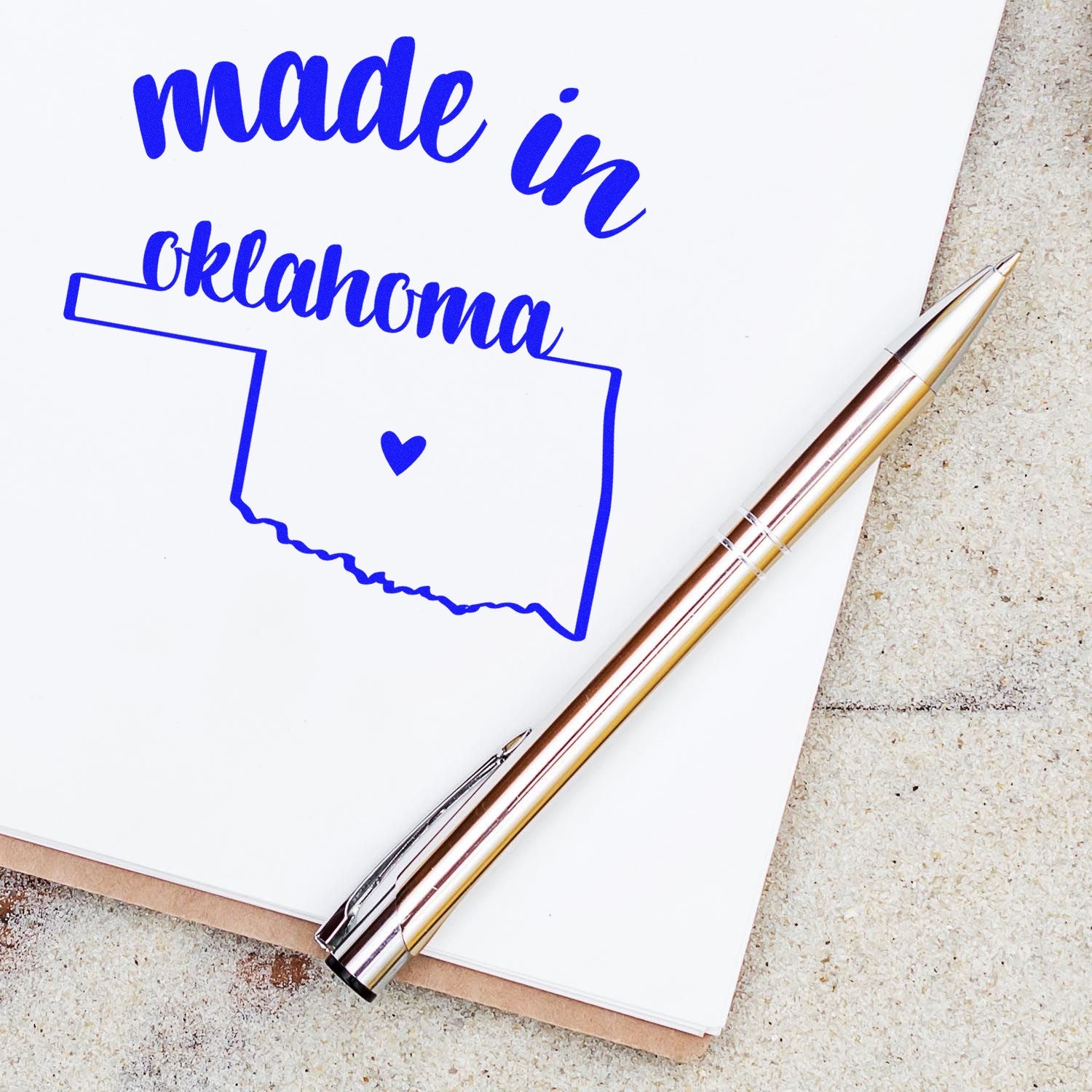 Self-Inking Handmade with Love in Oklahoma Stamp on paper, featuring a blue outline of Oklahoma with a heart, alongside a gold pen. Perfect for crafts and personalized projects.