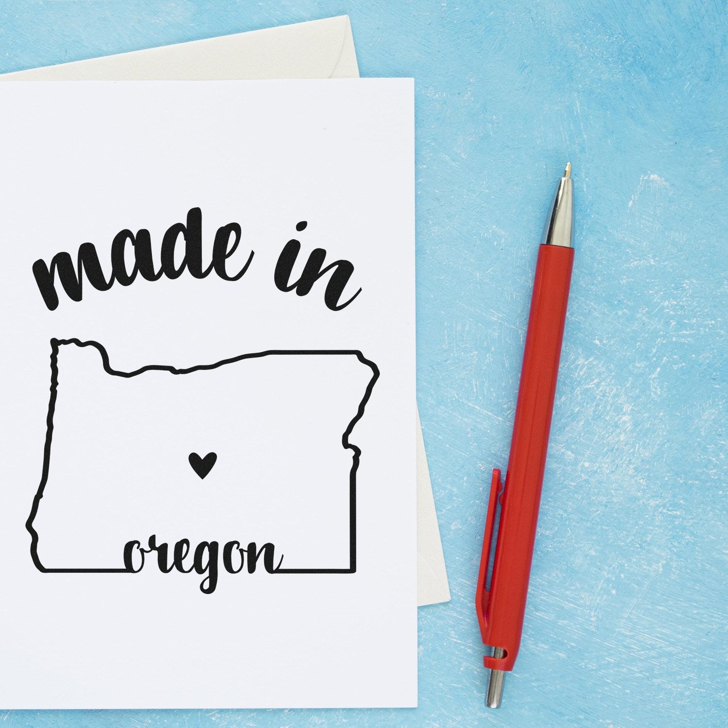 Made in Oregon Stamp Pre-Inked, featuring a black outline of Oregon with a heart, on a white card with a red pen on a blue background.