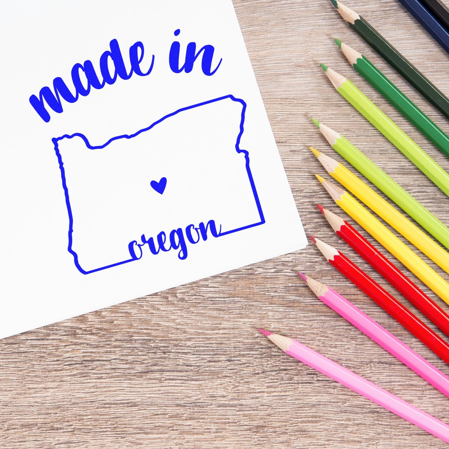 Image of the Slim Pre-Inked Stamp Oregon Made in Stamp, featuring a blue outline of Oregon with made in oregon text, next to colorful pencils on a wooden surface.